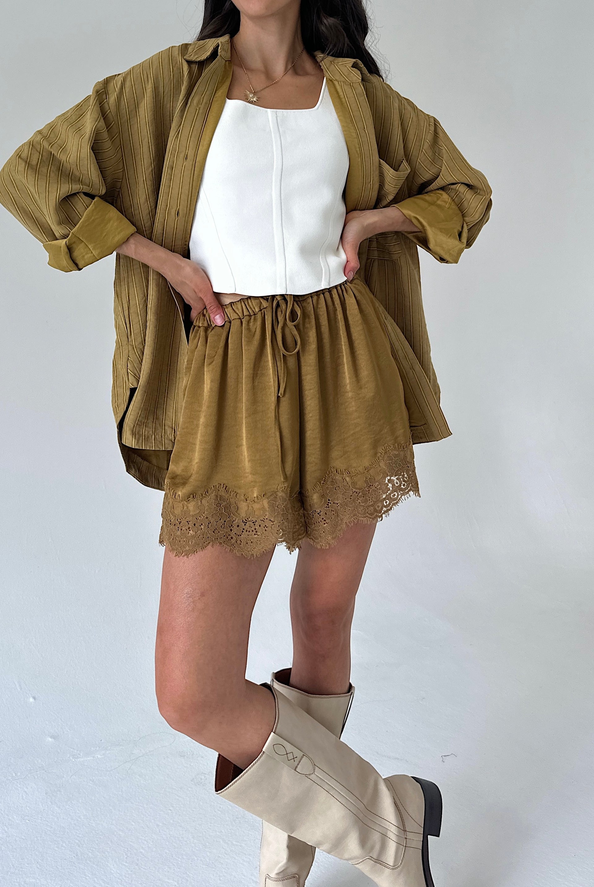 A model wearing a matching two-piece set consisting of a cropped, long-sleeve olive-brown shirt and matching drawstring shorts with lace trim, styled over a white tank top and cream-colored knee-high boots.