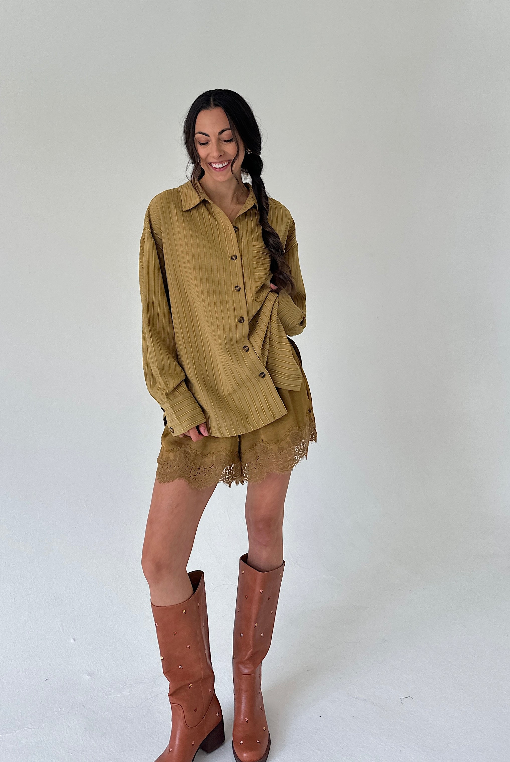 A woman modeling a matching mustard button-up shirt and lace shorts set, paired with brown leather knee-high boots, against a plain light gray studio background.