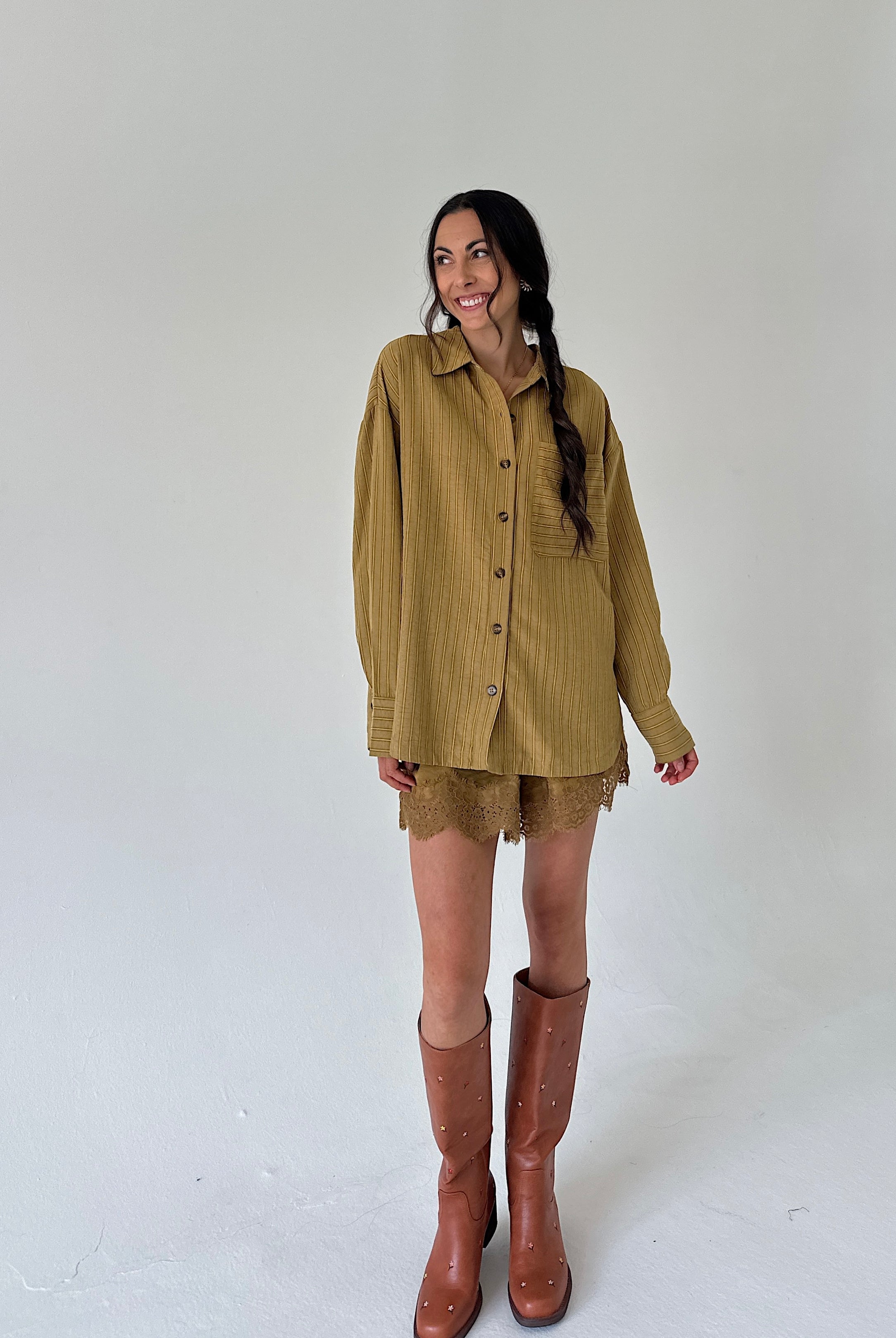 A woman modeling a loose olive-brown button-up shirt with matching lace shorts and brown knee-high boots, standing against a plain light gray studio background.