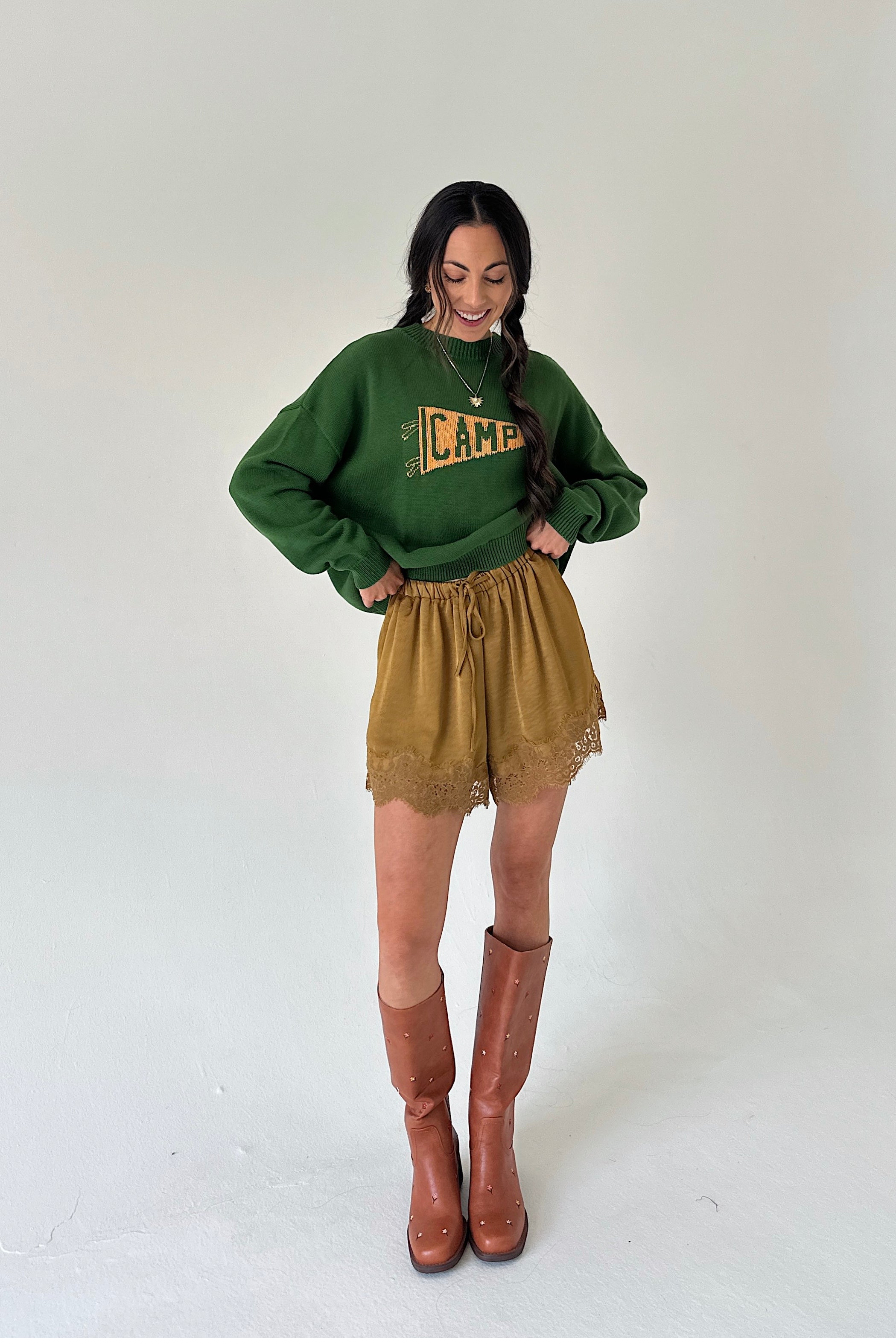 A woman modeling a dark green crewneck sweater with a yellow 'CAMP' graphic, paired with mustard lace-trim shorts and brown knee-high boots, against a plain light gray studio background.