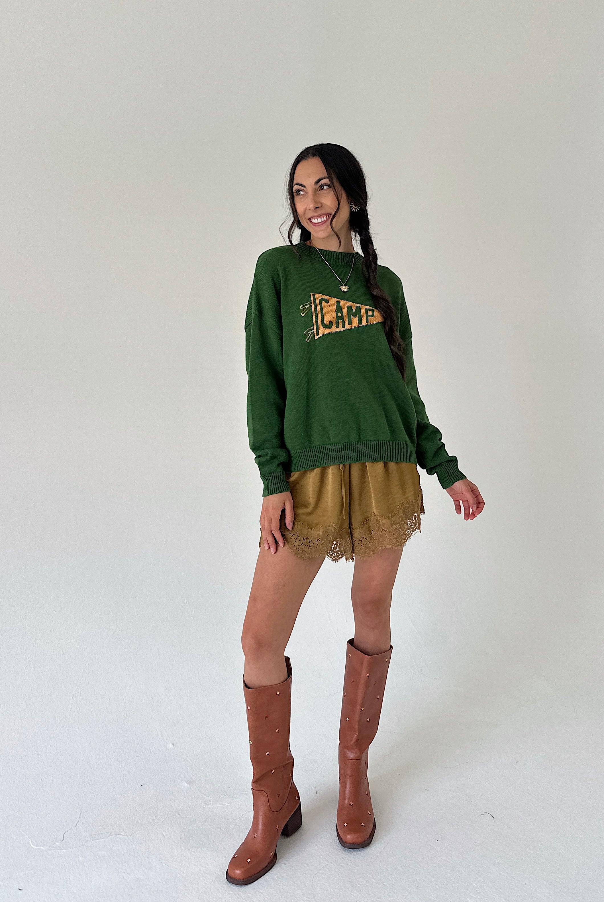 A woman modeling a dark green crewneck sweater with a distressed yellow 'CAMP' graphic, paired with mustard lace hem shorts and brown knee-high boots, against a plain off-white studio background.