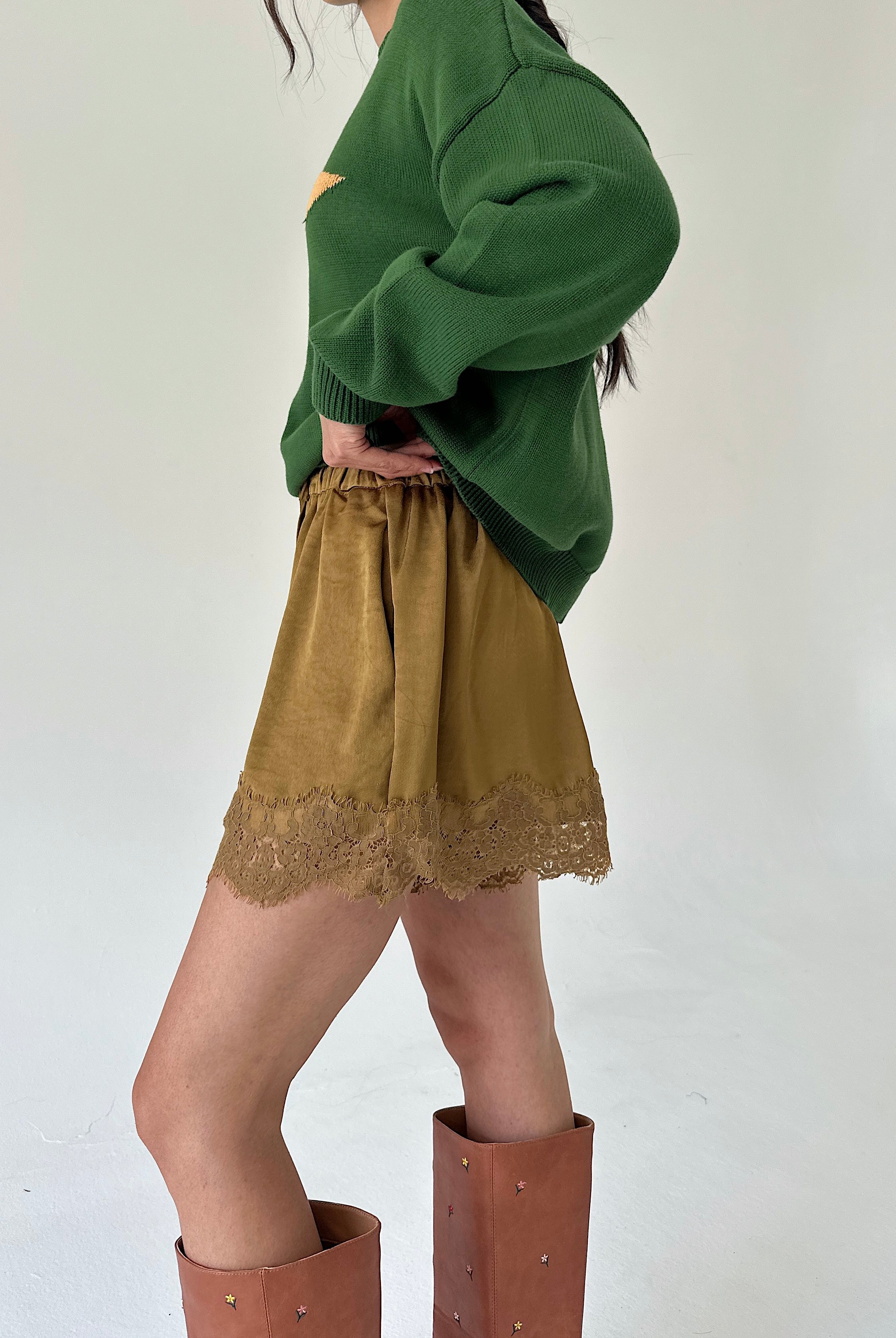 A person wearing an oversized green sweater, mustard shorts with a lace hem, and tall brown boots, photographed against a plain light background.