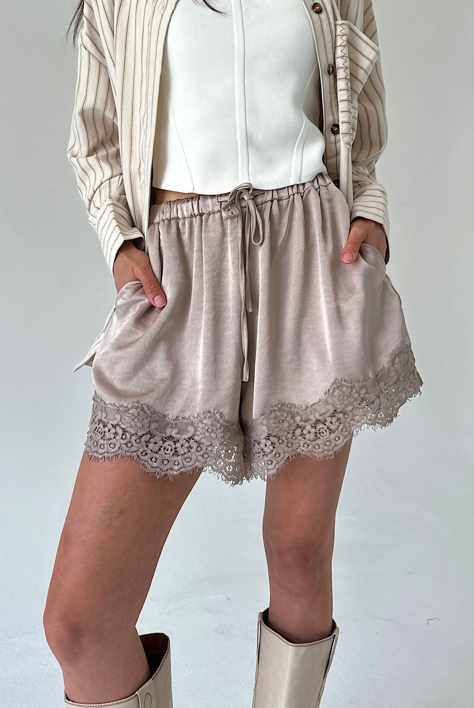 A person wearing light taupe satin shorts with a lace hem, styled with a white top, a beige striped cardigan, and beige boots against a plain light gray studio background.