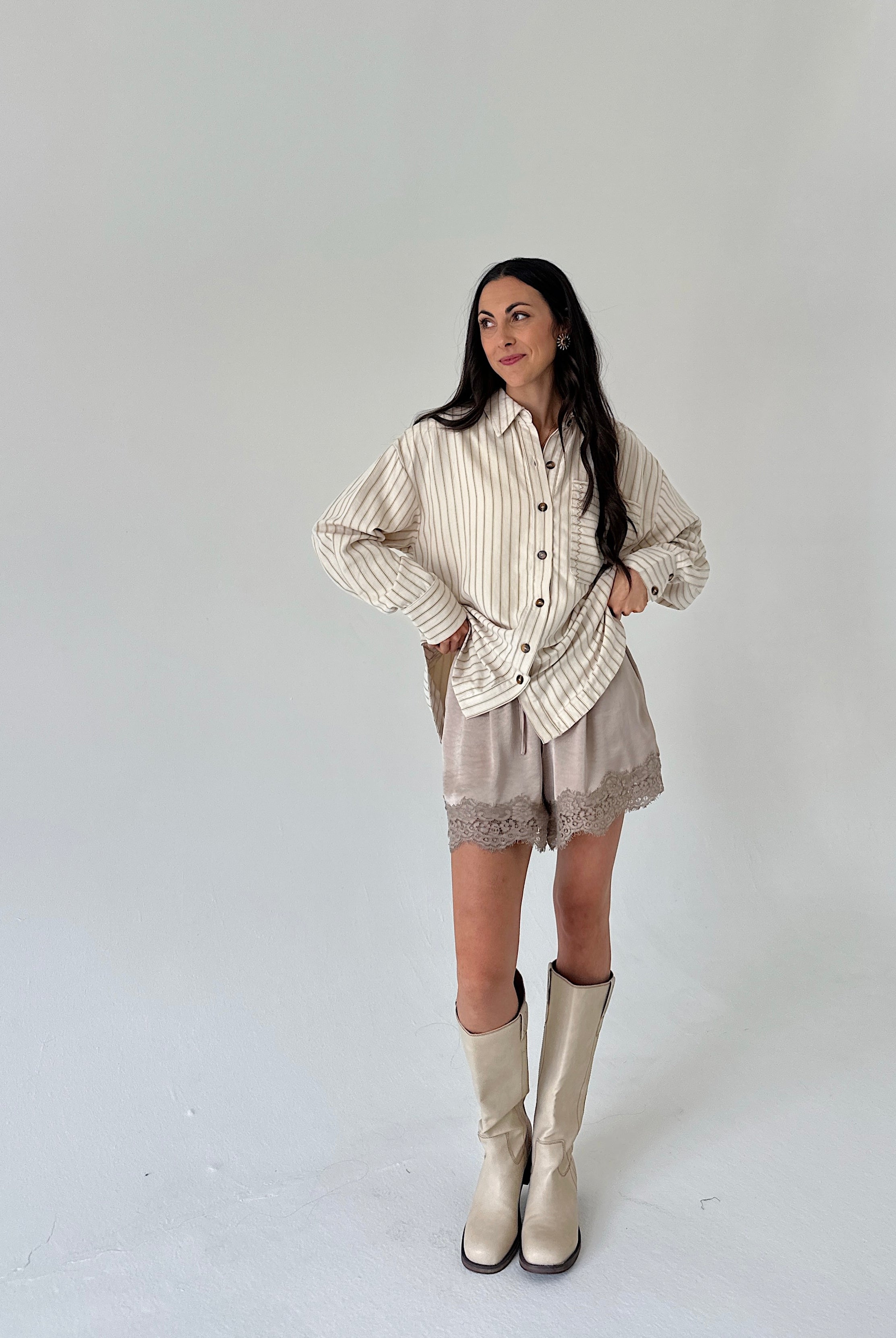 A woman modeling a cream and beige vertically striped button-up shirt, matching beige shorts with lace trim, and beige knee-high boots against a plain light gray studio background.