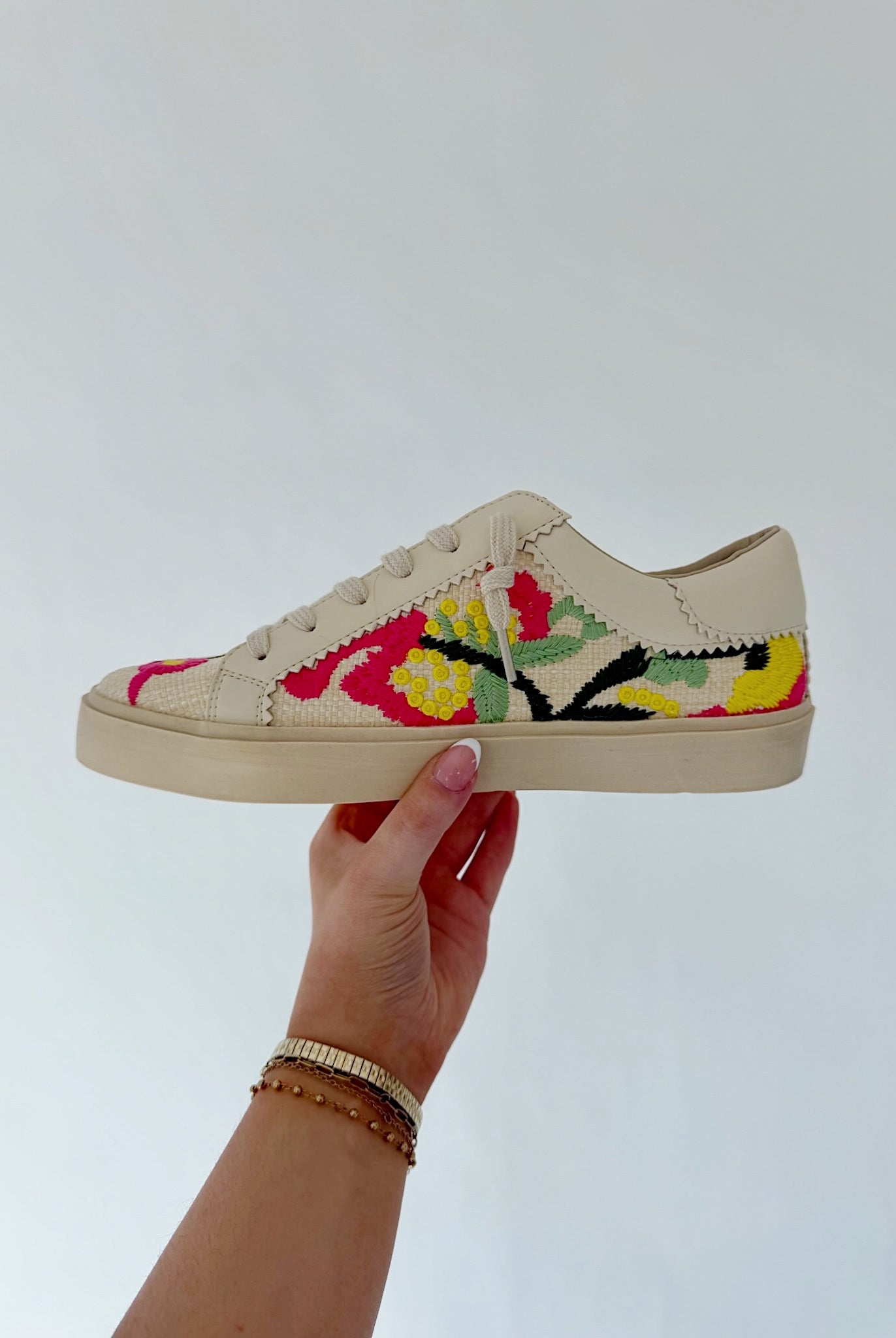 Beige sneaker with colorful floral embroidery held by a hand against a light gray background