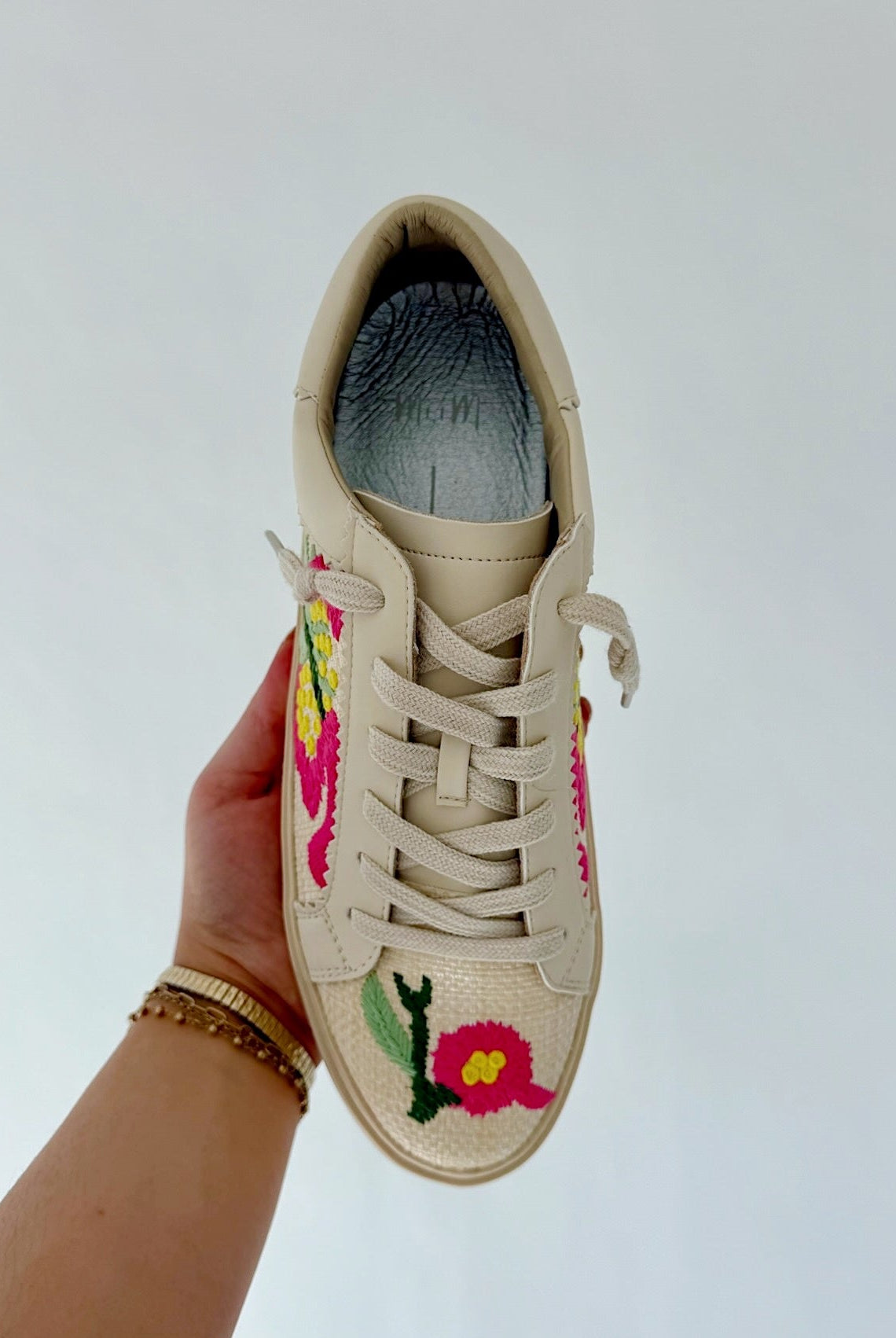 Beige sneaker with floral embroidery held by a hand against a light gray background
