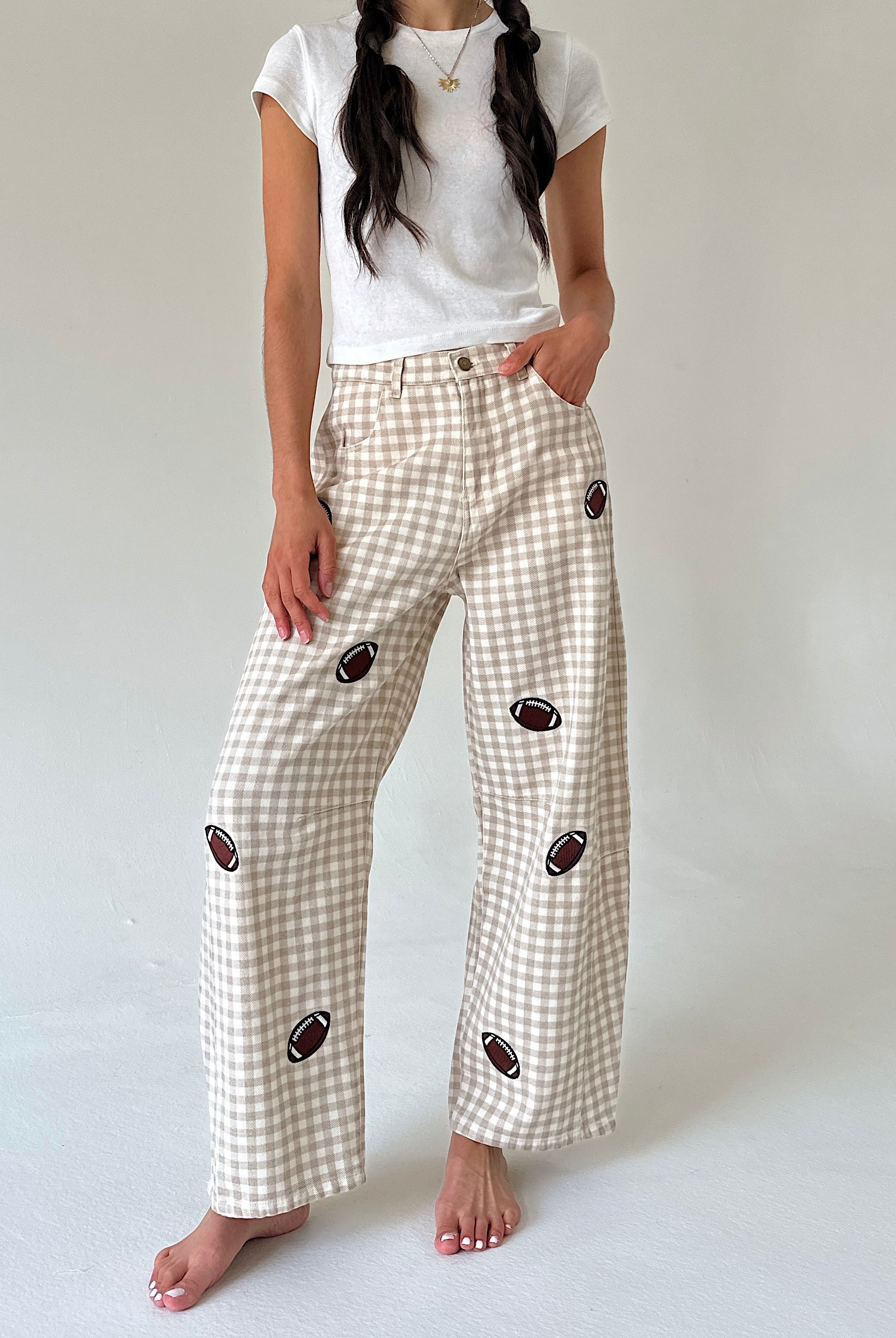 A model wearing beige and white gingham wide-leg pants with embroidered football motifs, paired with a white t-shirt, standing against a neutral light gray background.