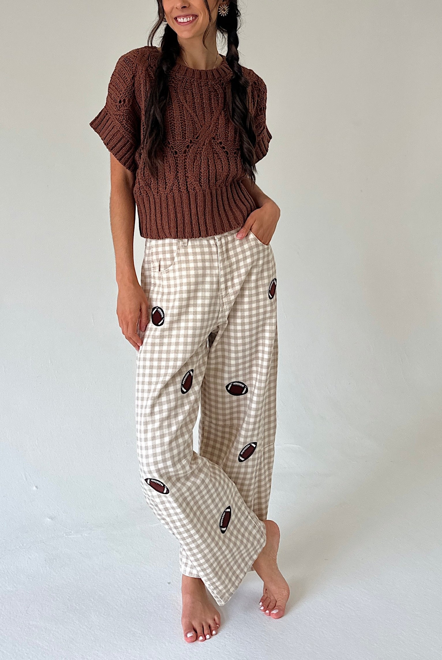 A woman modeling a brown short-sleeve knit sweater and beige gingham pants with football prints, standing against a plain light gray studio background.