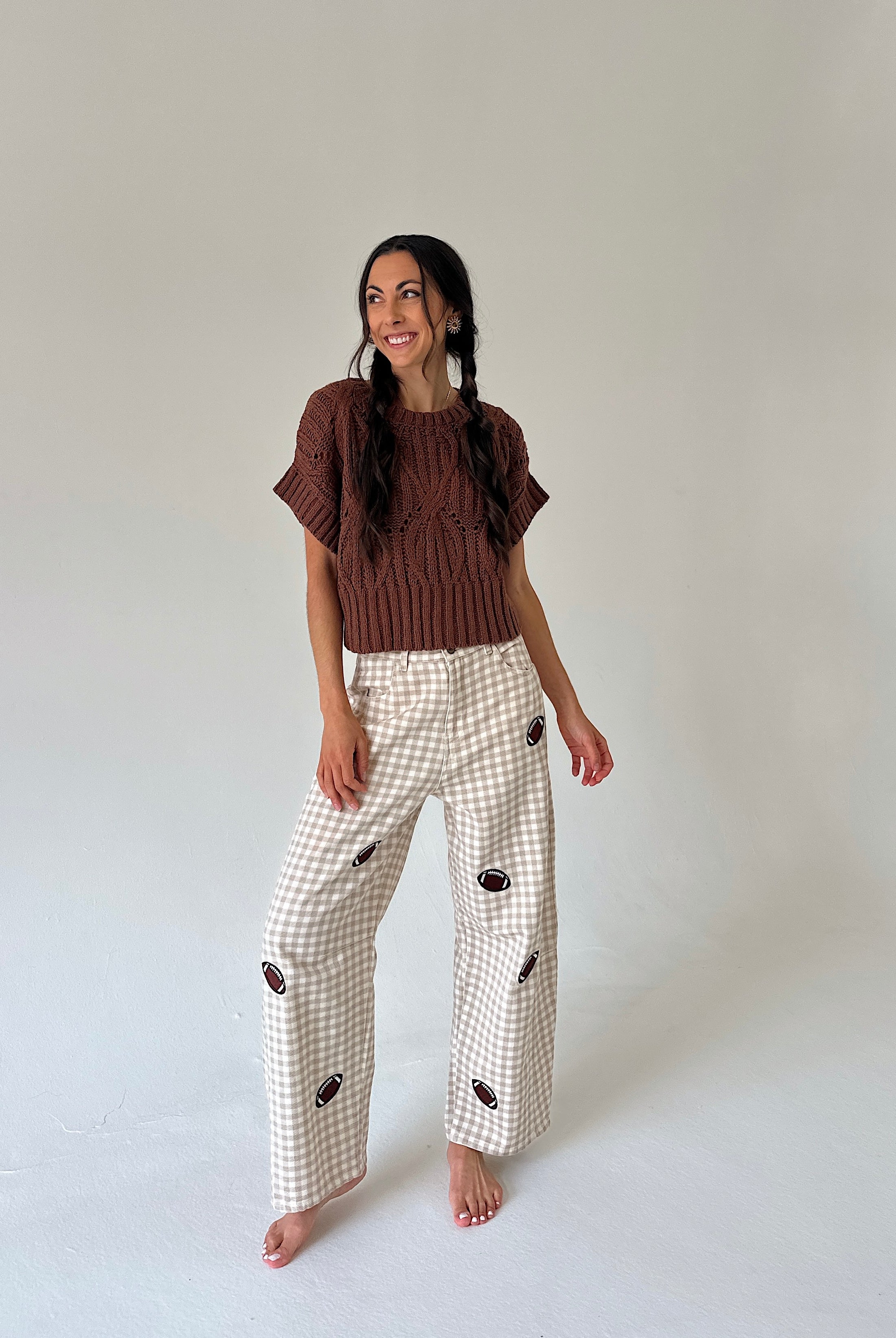 A woman stands barefoot against a plain light gray studio background, wearing a brown short-sleeve knit top and cream gingham wide-leg pants with small football patches.