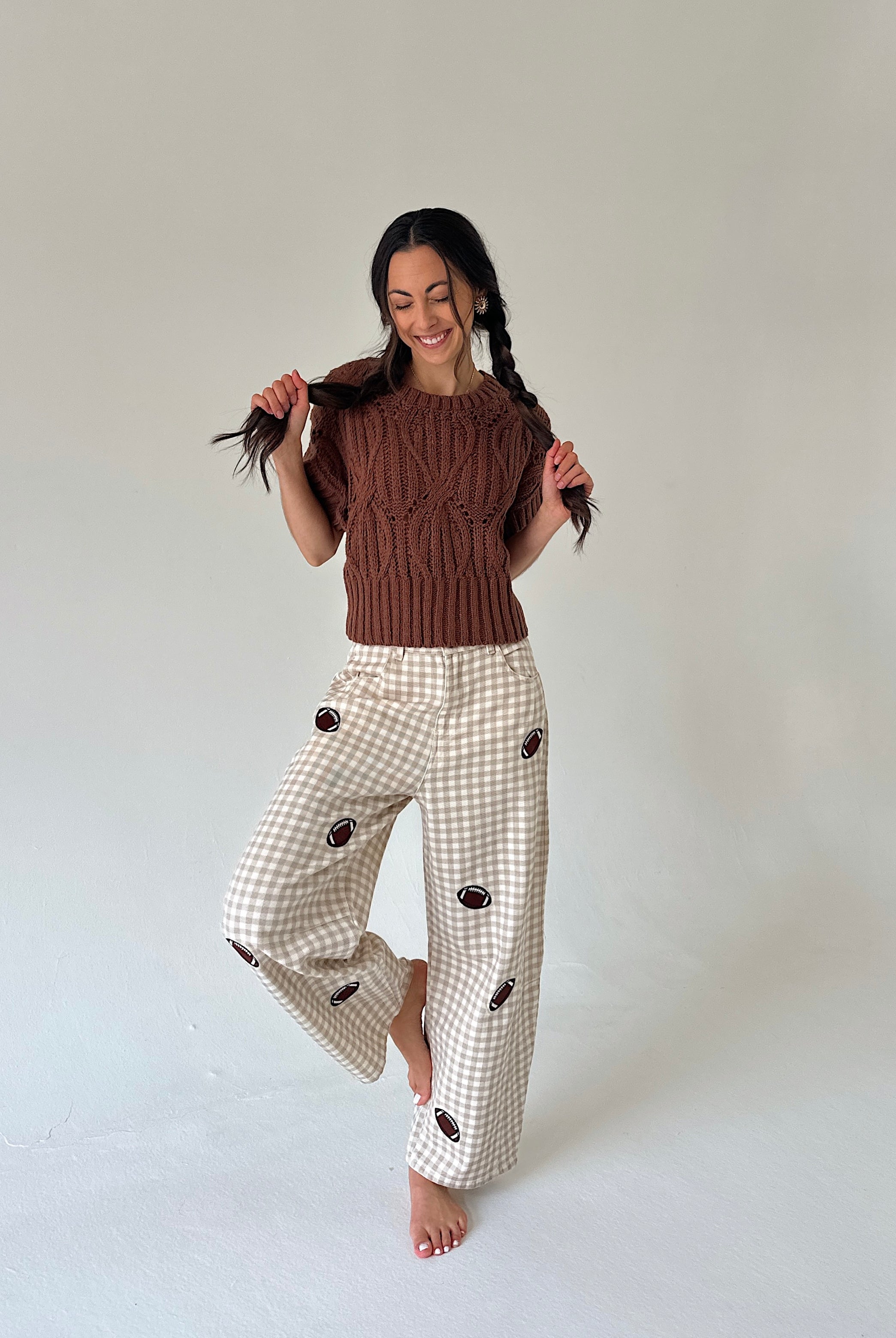 A woman posing in a studio setting, wearing a brown cable-knit short-sleeve top and cream gingham wide-leg pants with dark brown embroidered details.