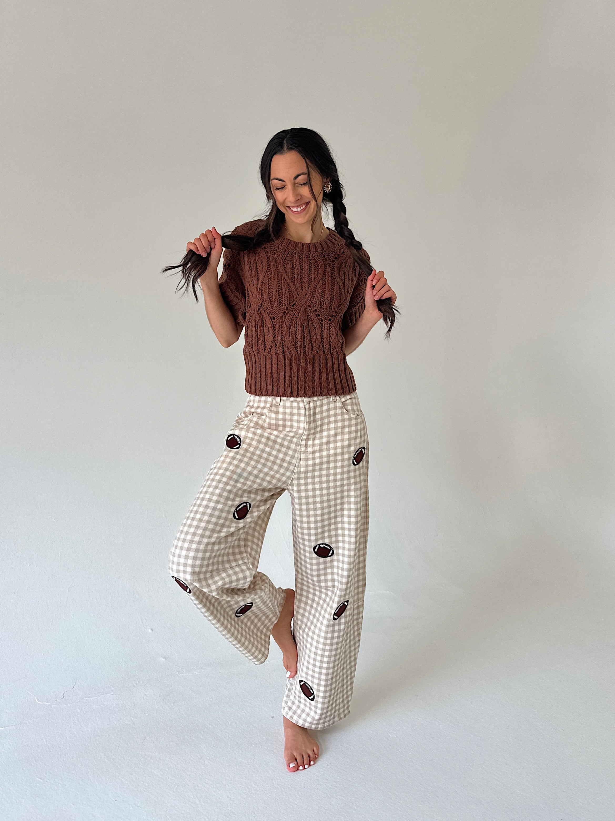 A woman posing in a studio setting, wearing a brown cable-knit short-sleeve top and cream gingham wide-leg pants with dark brown embroidered details.