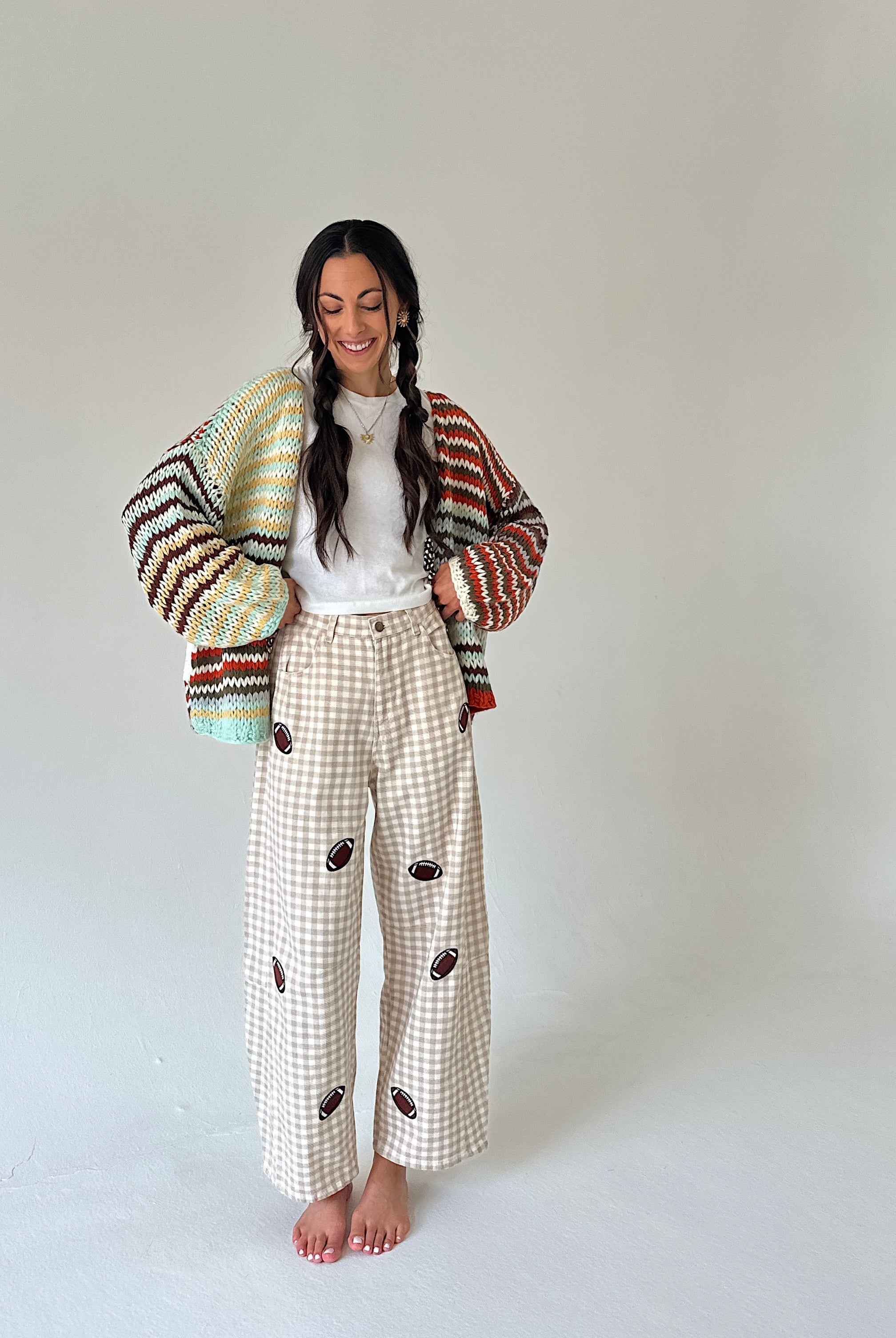 A woman stands barefoot against a plain light gray studio background, wearing a multicolor knit cardigan draped off her shoulders, a white top, and cream gingham wide-leg pants with brown embroidered details.