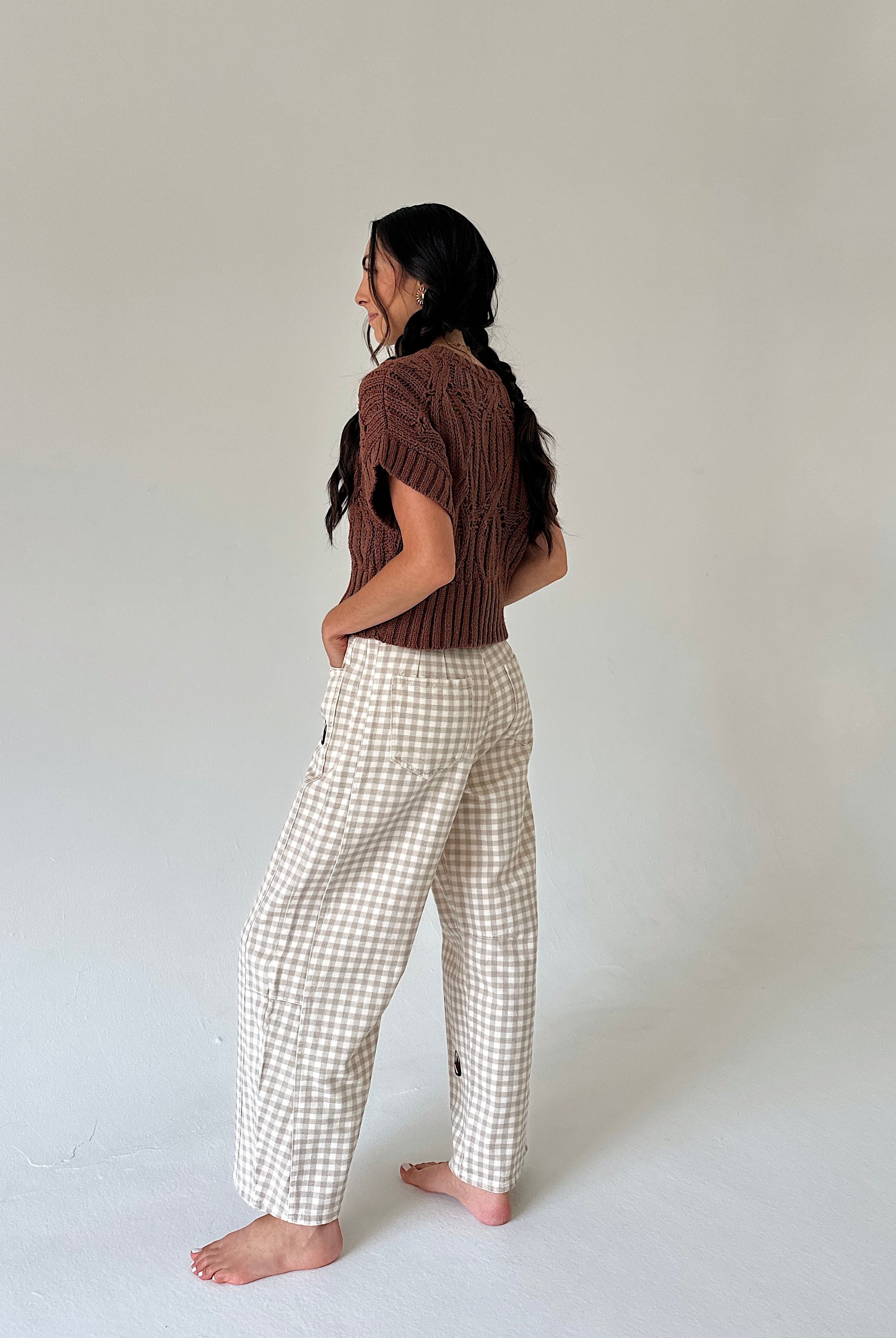 A woman stands barefoot in profile wearing a brown short-sleeve knit top and cream-and-white gingham wide-leg pants against a plain light gray studio background.