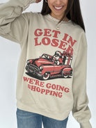 Woman wearing a sand sweatshirt with a car graphic that has Santa inside with words surrounding that says "Get in loser we're going shopping"