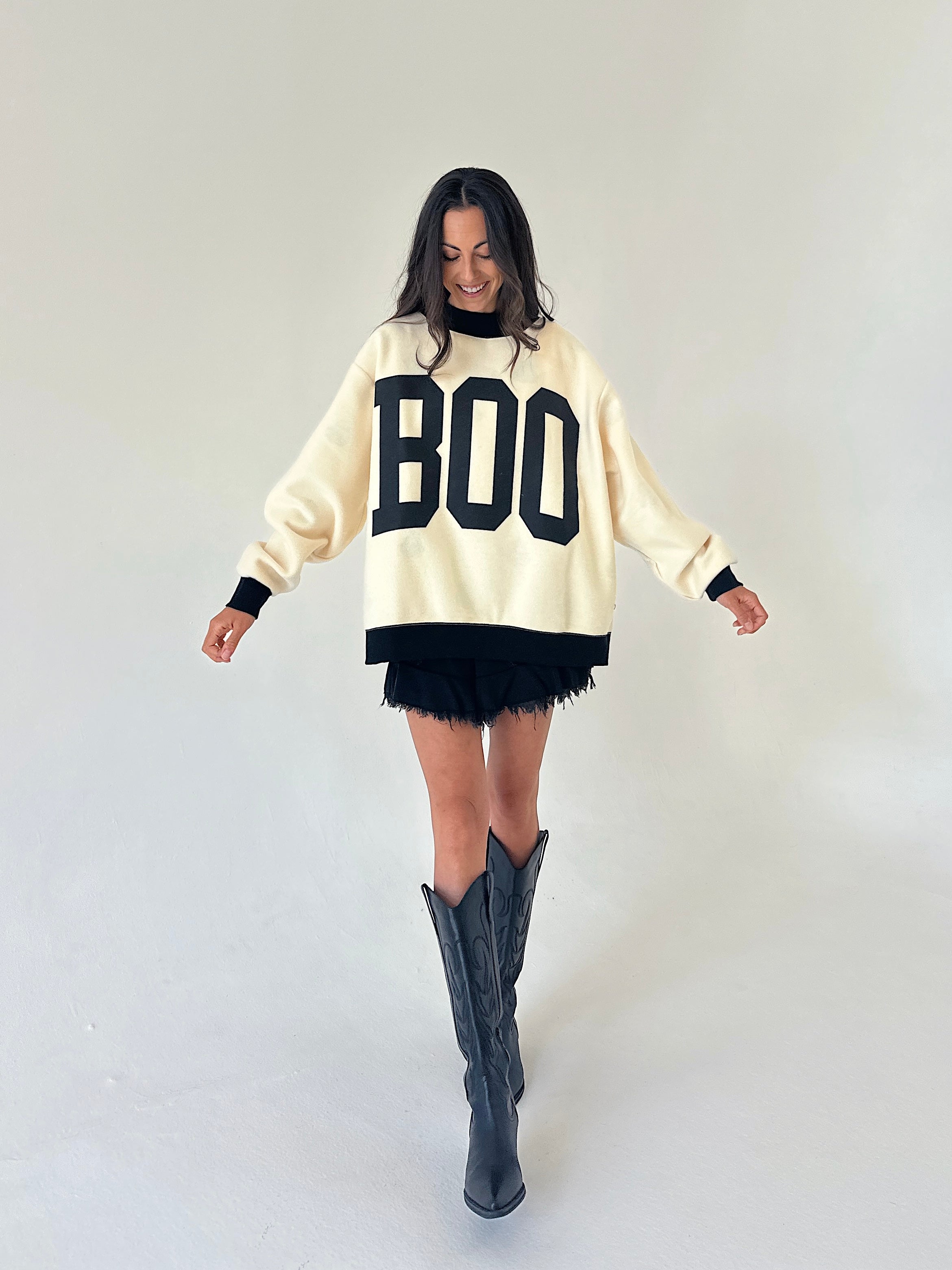 A woman modeling a cream-colored knit sweater with 'BOO' printed in black varsity letters across the chest, paired with a black fringed mini skirt and black cowboy boots, set against a plain light gray studio background.