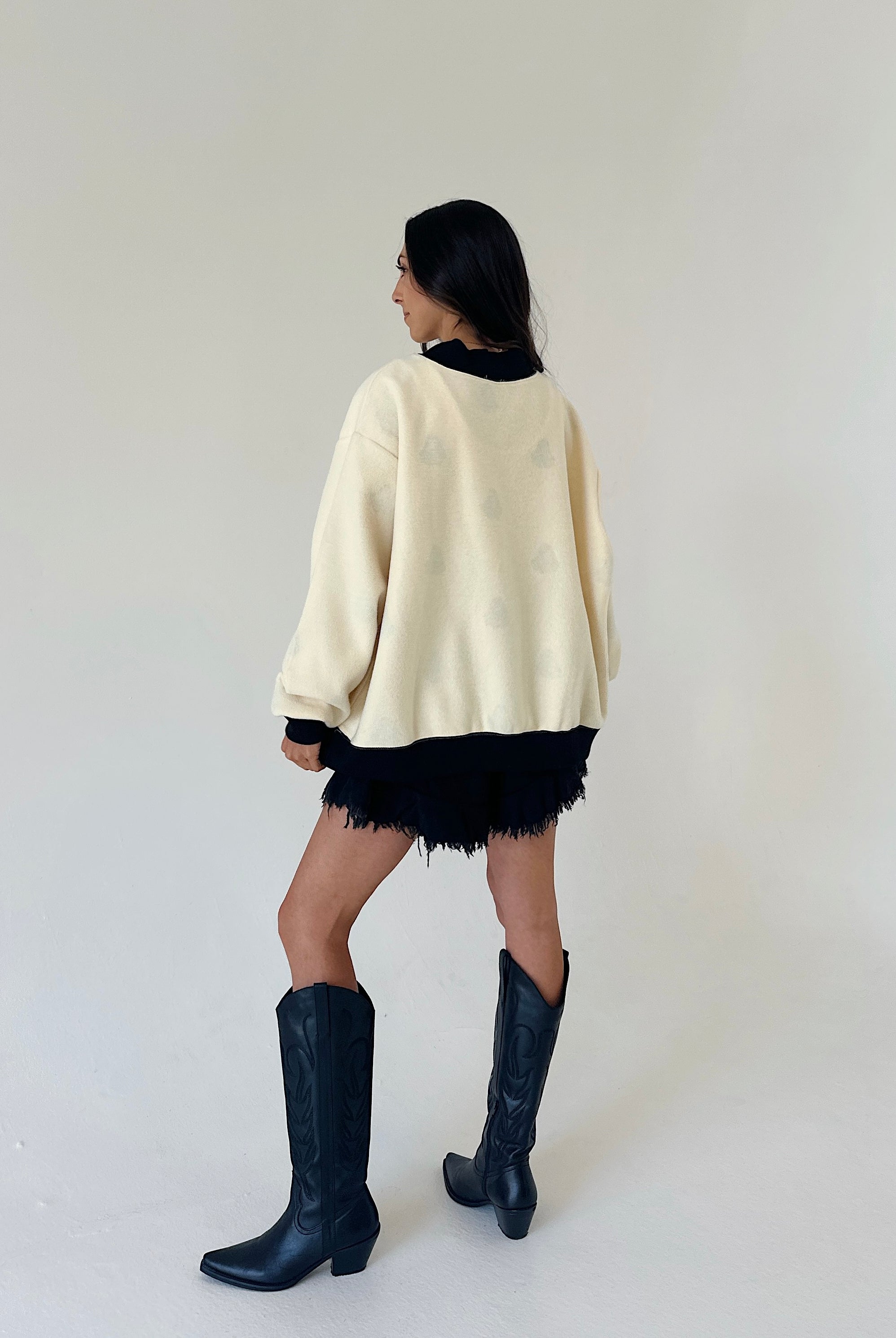 A woman is shown from behind wearing a cream-colored oversized sweater, a black fringed mini skirt, and black leather cowboy boots, set against a plain off-white studio background.