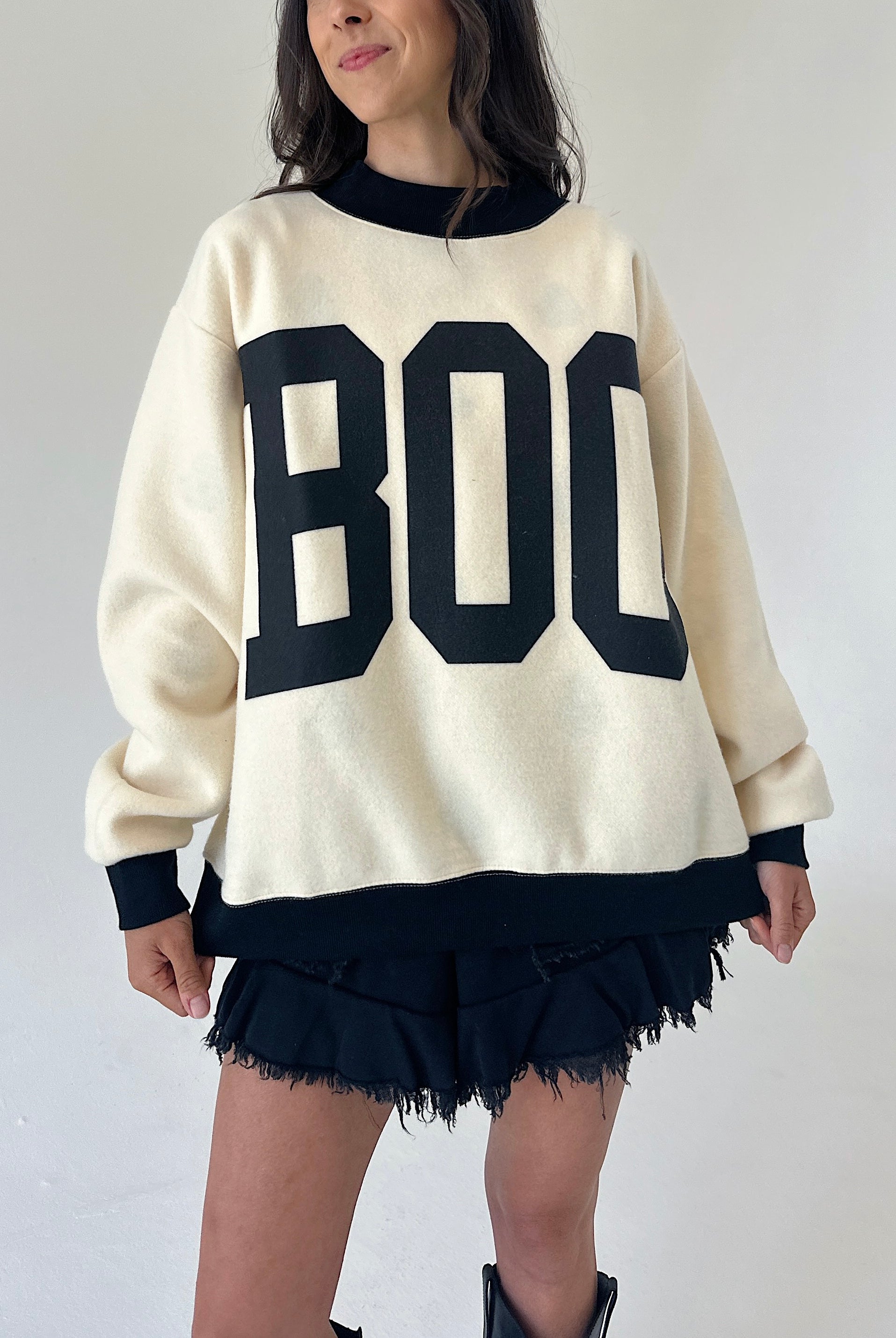 A woman wearing an oversized cream-colored sweater with large black block letters spelling 'BOO' across the front, paired with black frayed shorts and black boots, set against a plain light gray wall.
