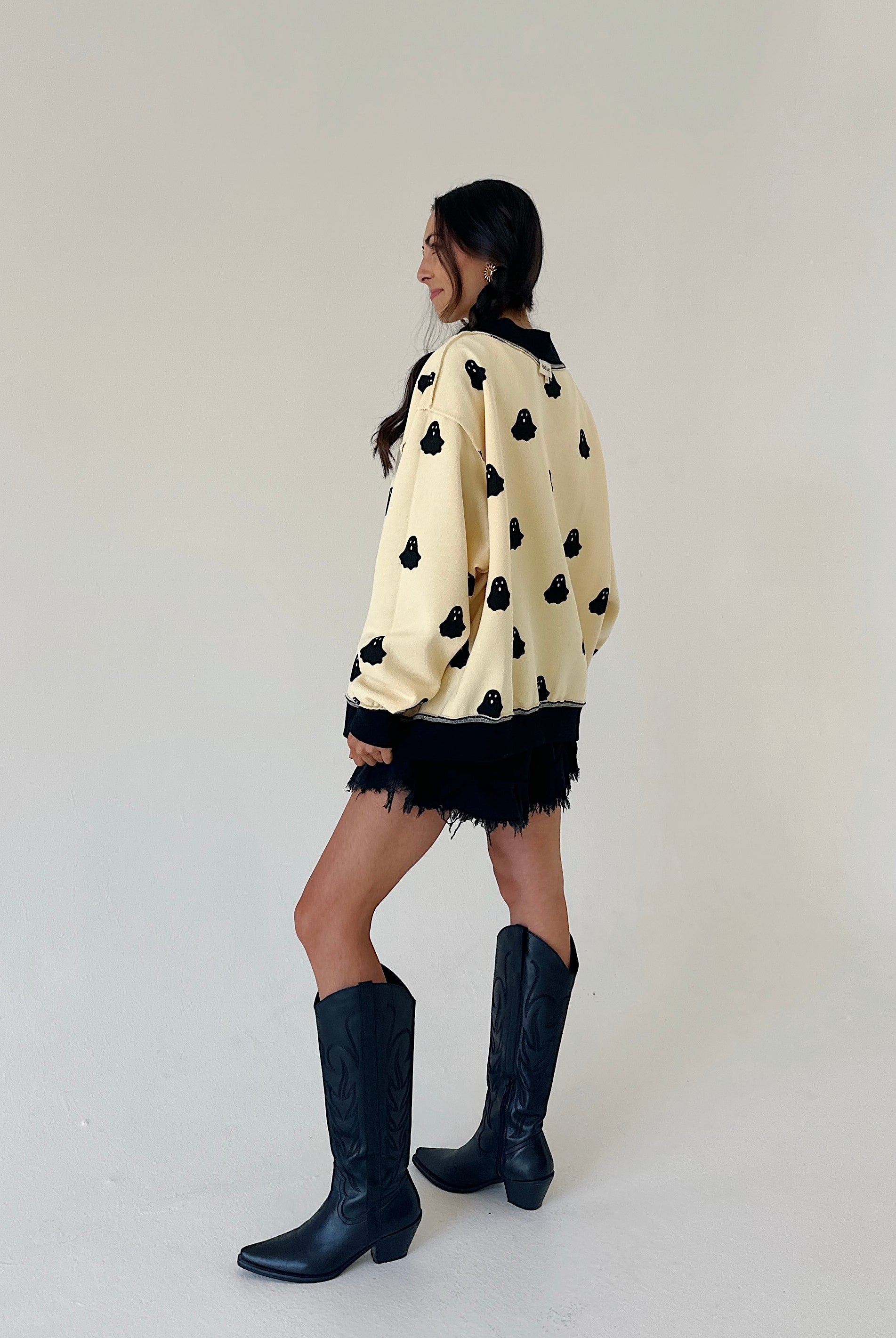 woman standing from a back side angle wearing a cream sweatshirt covered in black ghost graphics, wearing black shorts and tall black boots