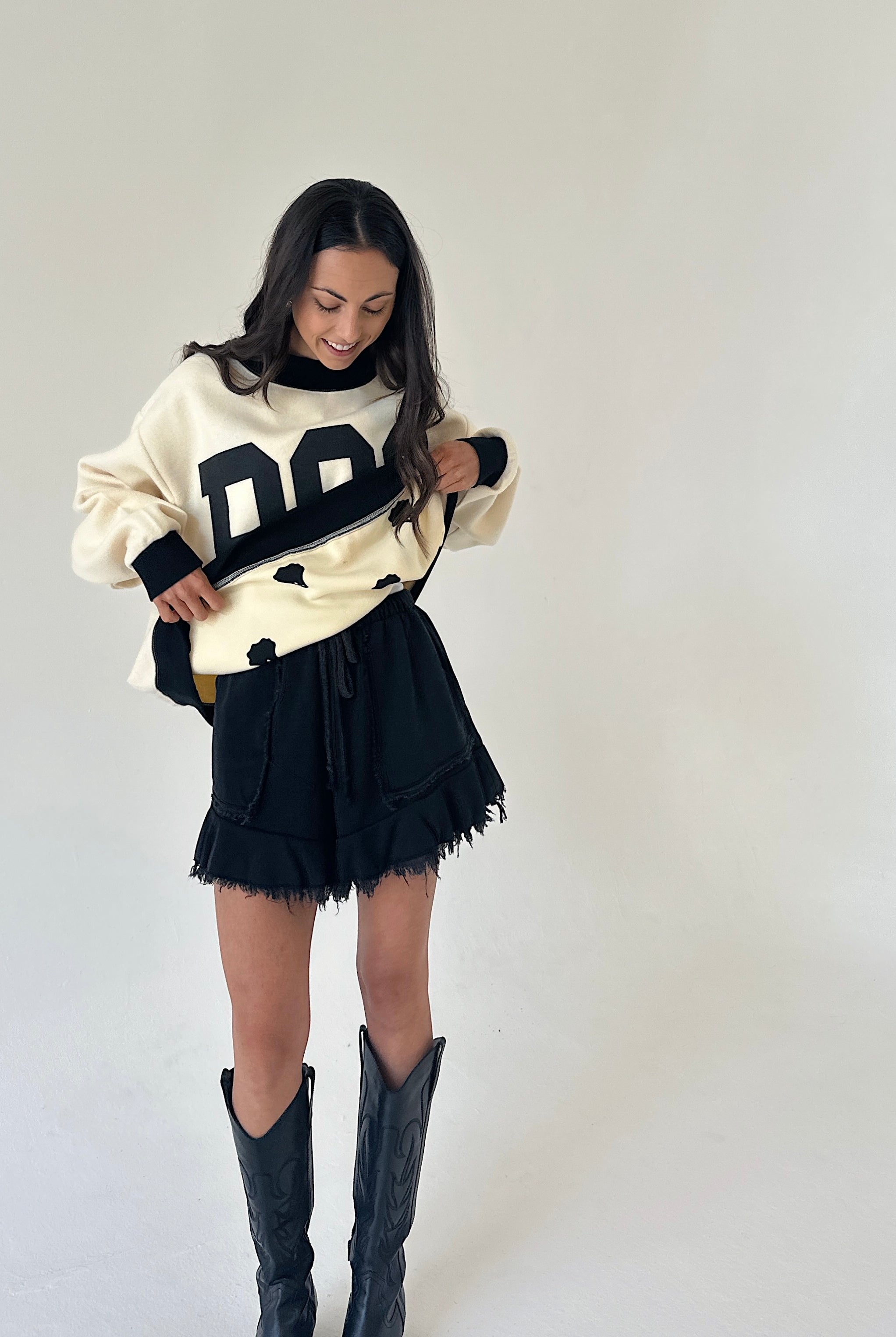 A woman modeling a cream and black reversible sweatshirt with large letter graphics, paired with black fringed shorts and black cowboy boots, against a plain off-white studio background.