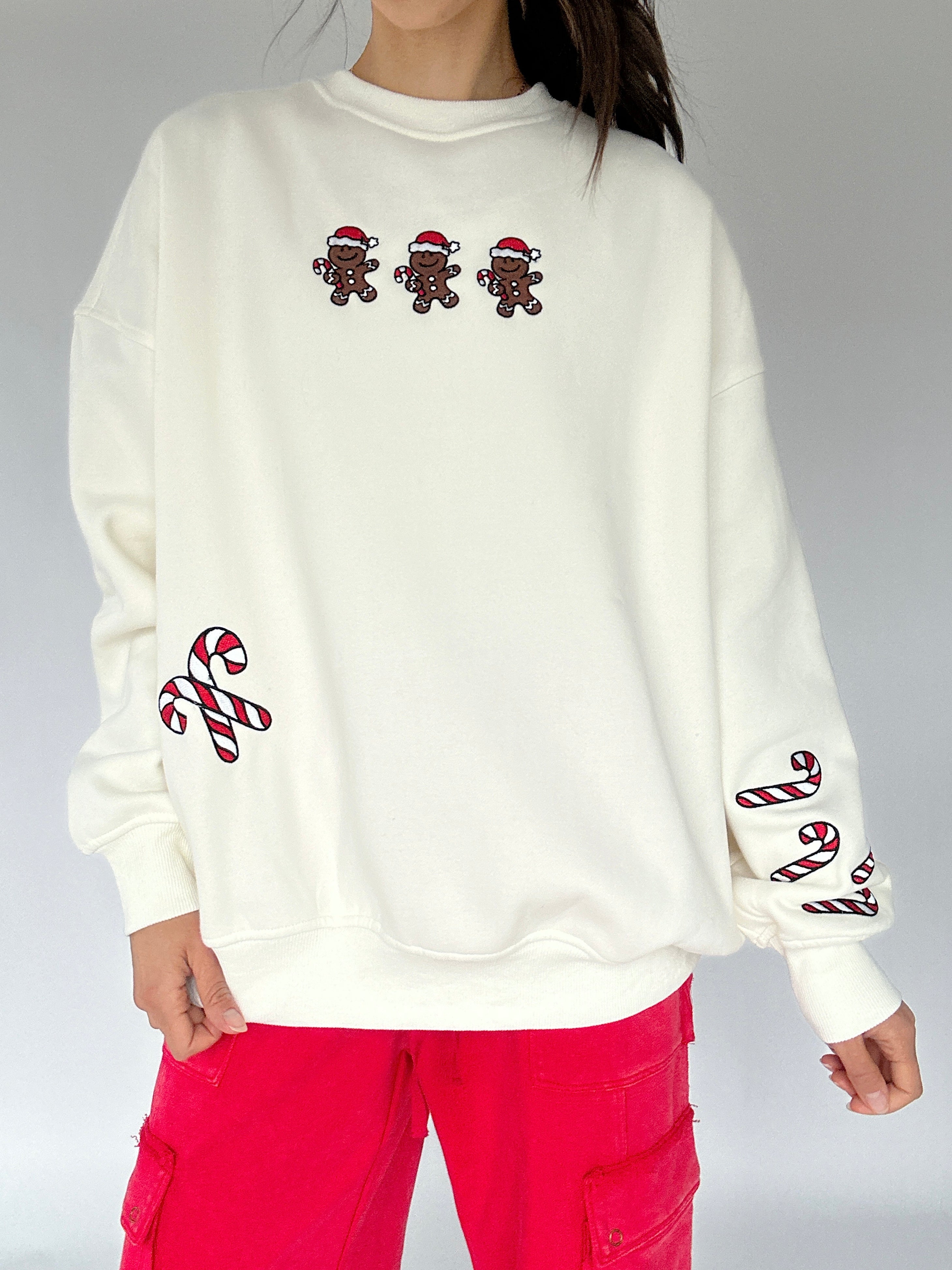 White sweatshirt with embroidered gingerbread men and candy canes, worn by a person with red pants.