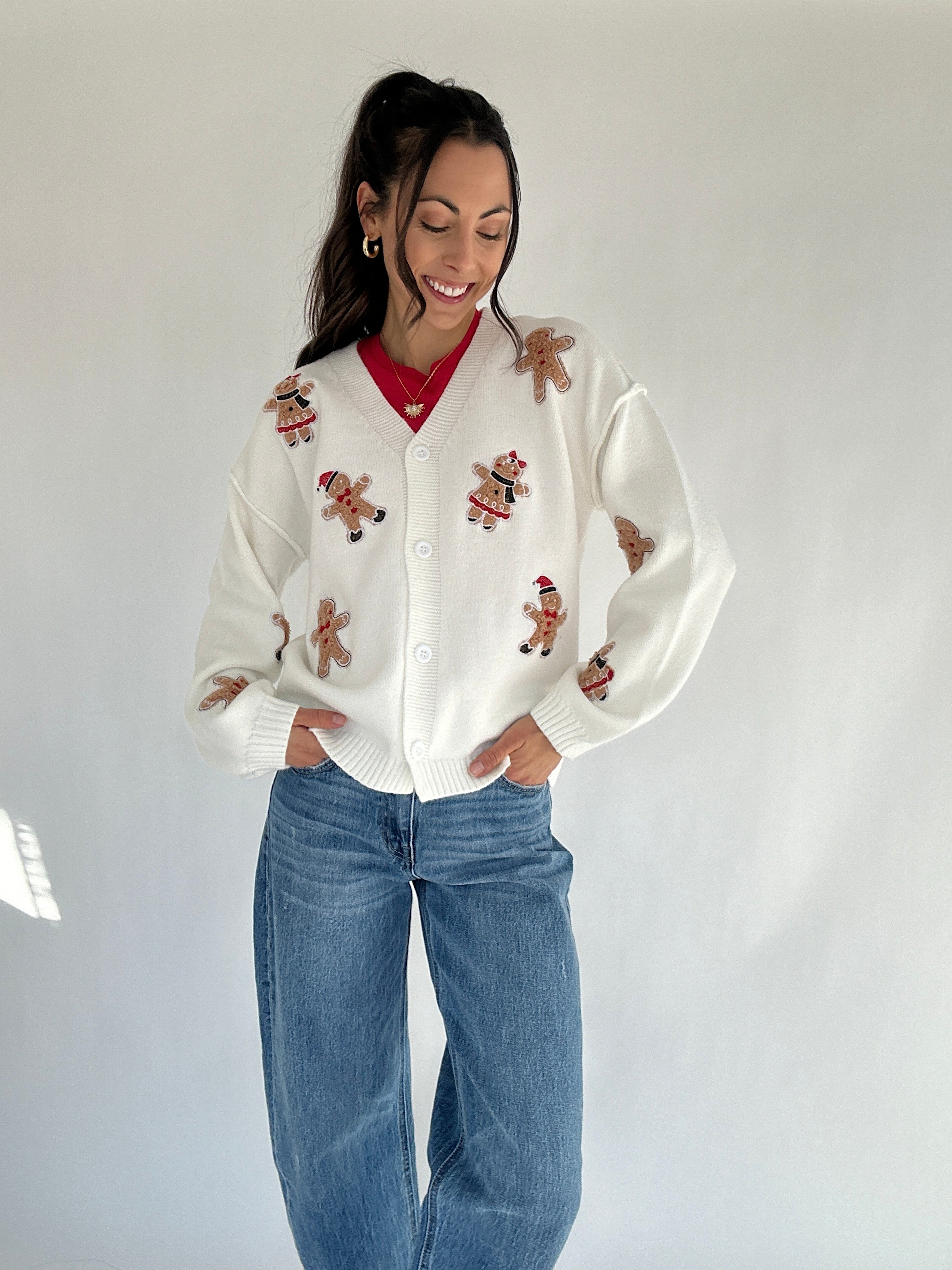 Woman wearing a white cardigan with gingerbread man patterns and blue jeans on a plain background