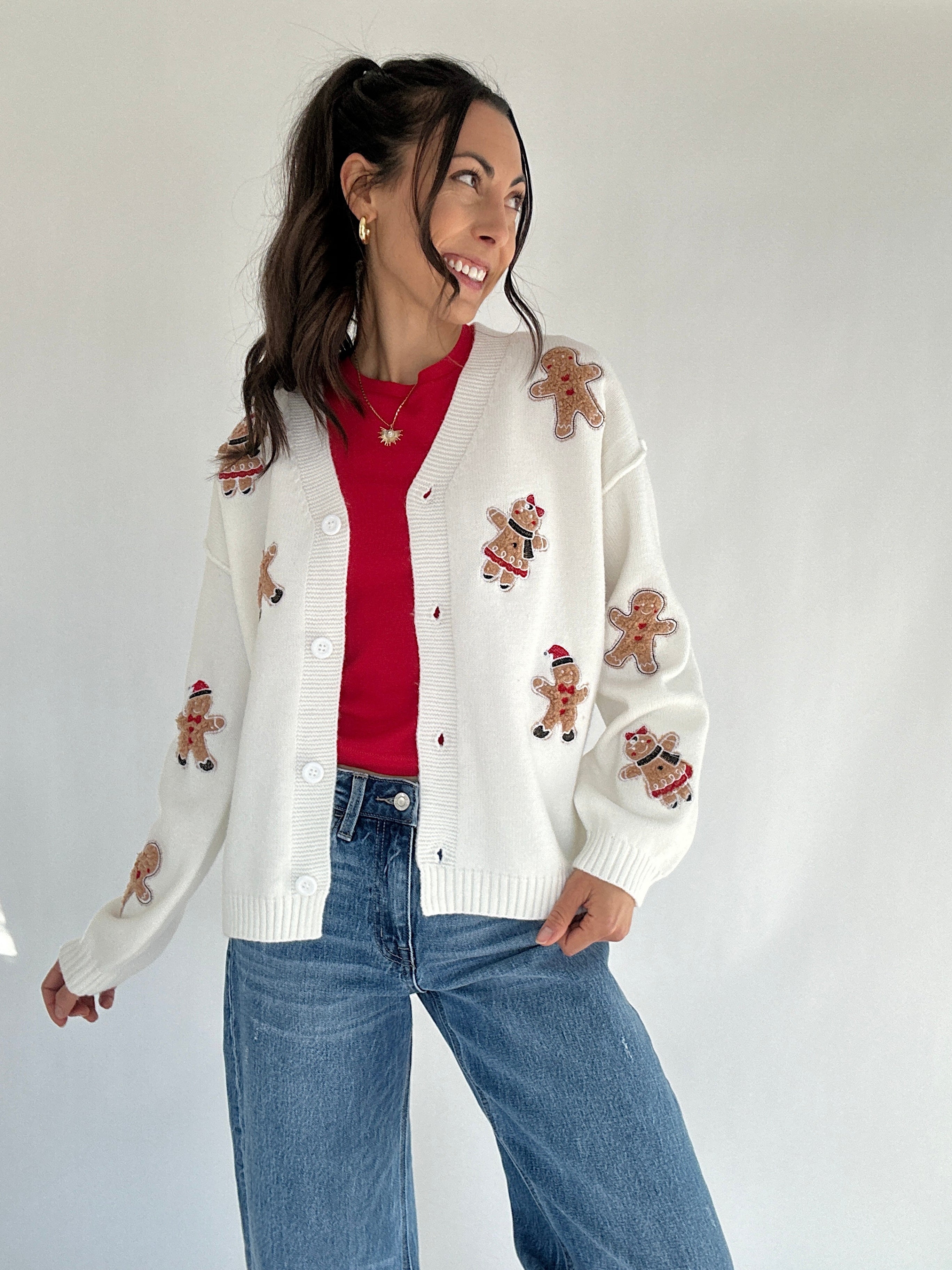 Woman wearing a white cardigan with gingerbread man patterns, red shirt, and blue jeans on a plain background