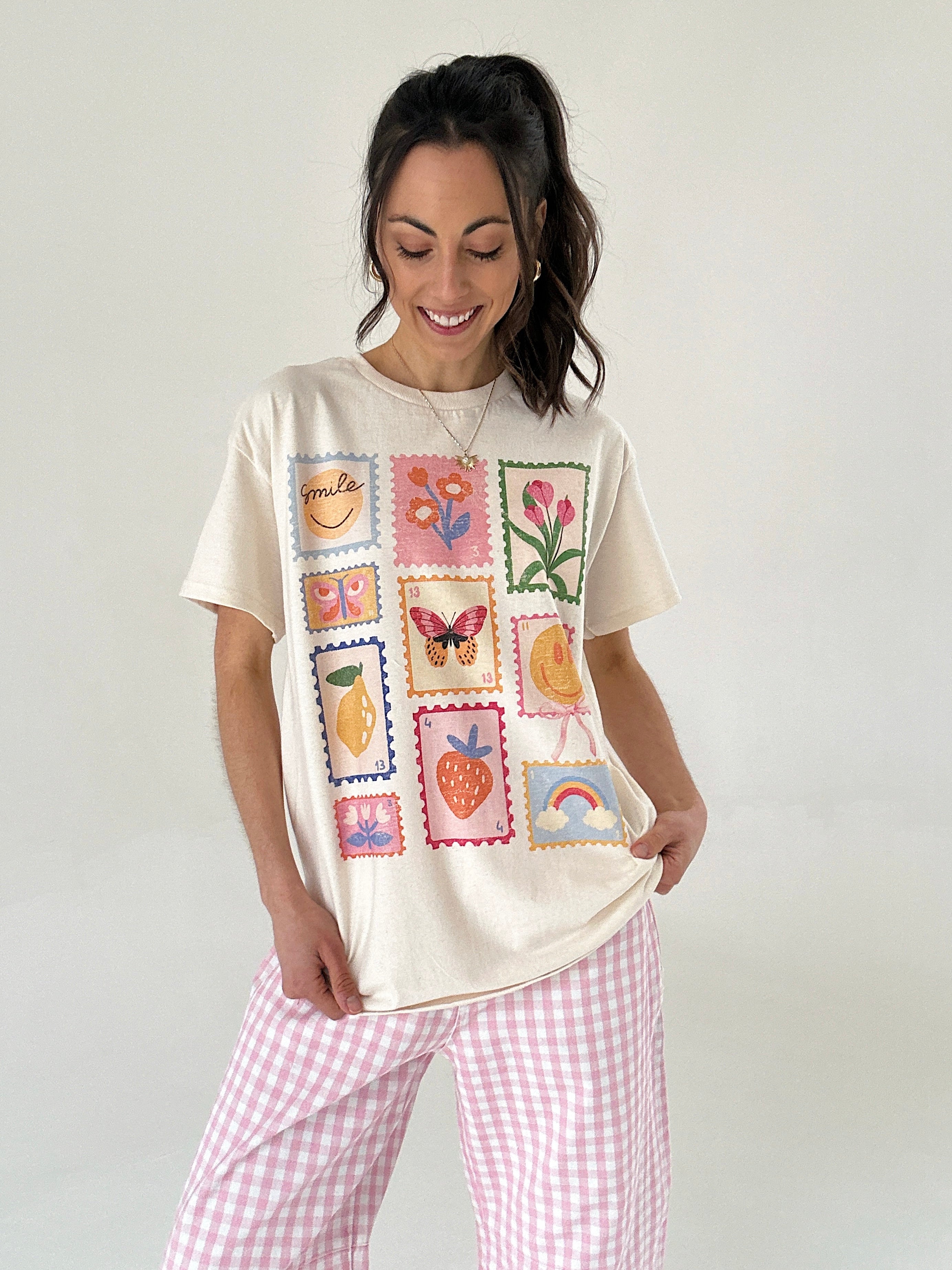 Woman wearing a cream t-shirt with colorful stamp designs and pink checkered pants on a white background