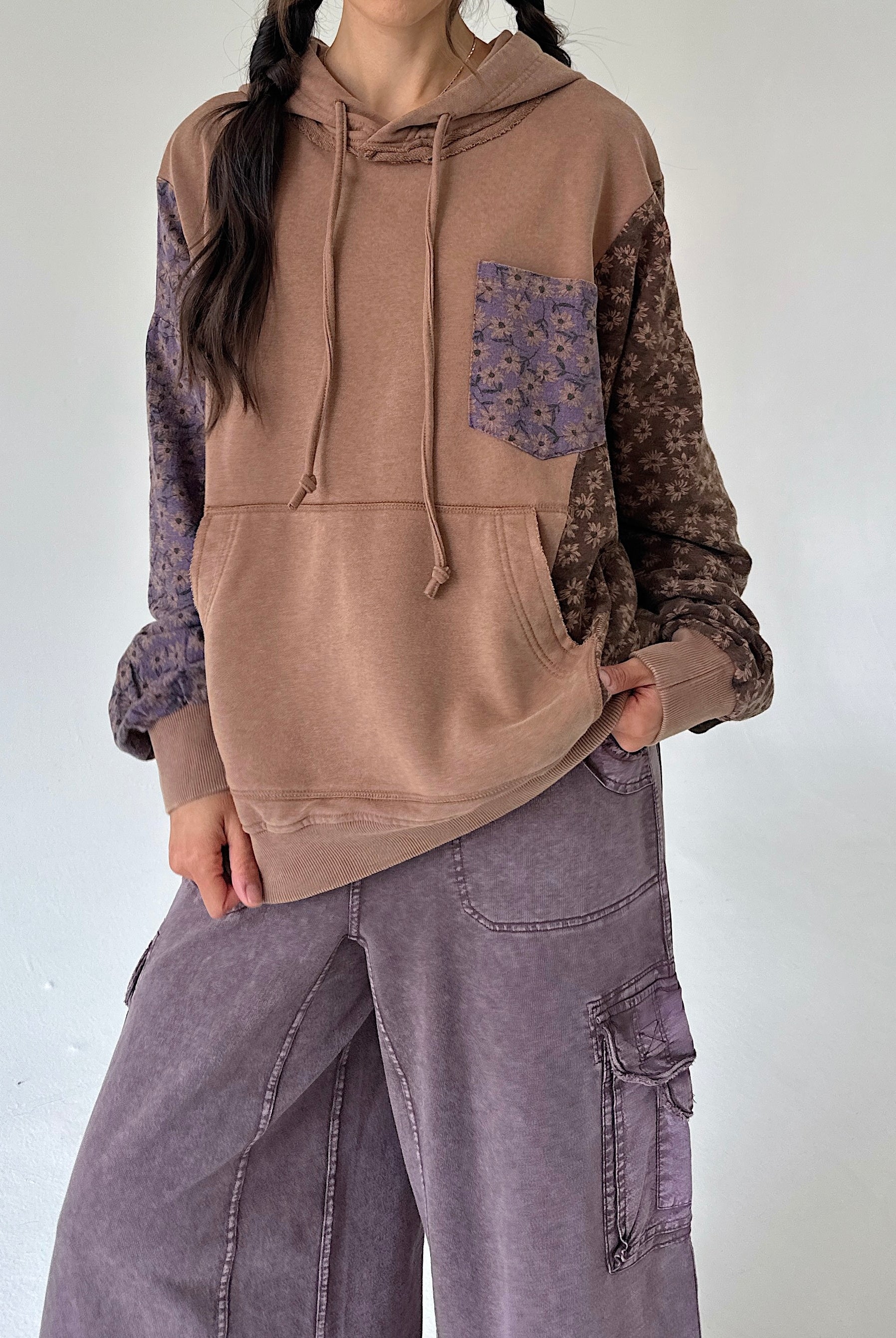 A person wearing a color-blocked hoodie with a solid tan front, patterned brown sleeves, and a purple floral chest pocket, paired with wide-leg mauve cargo pants against a plain light gray wall.