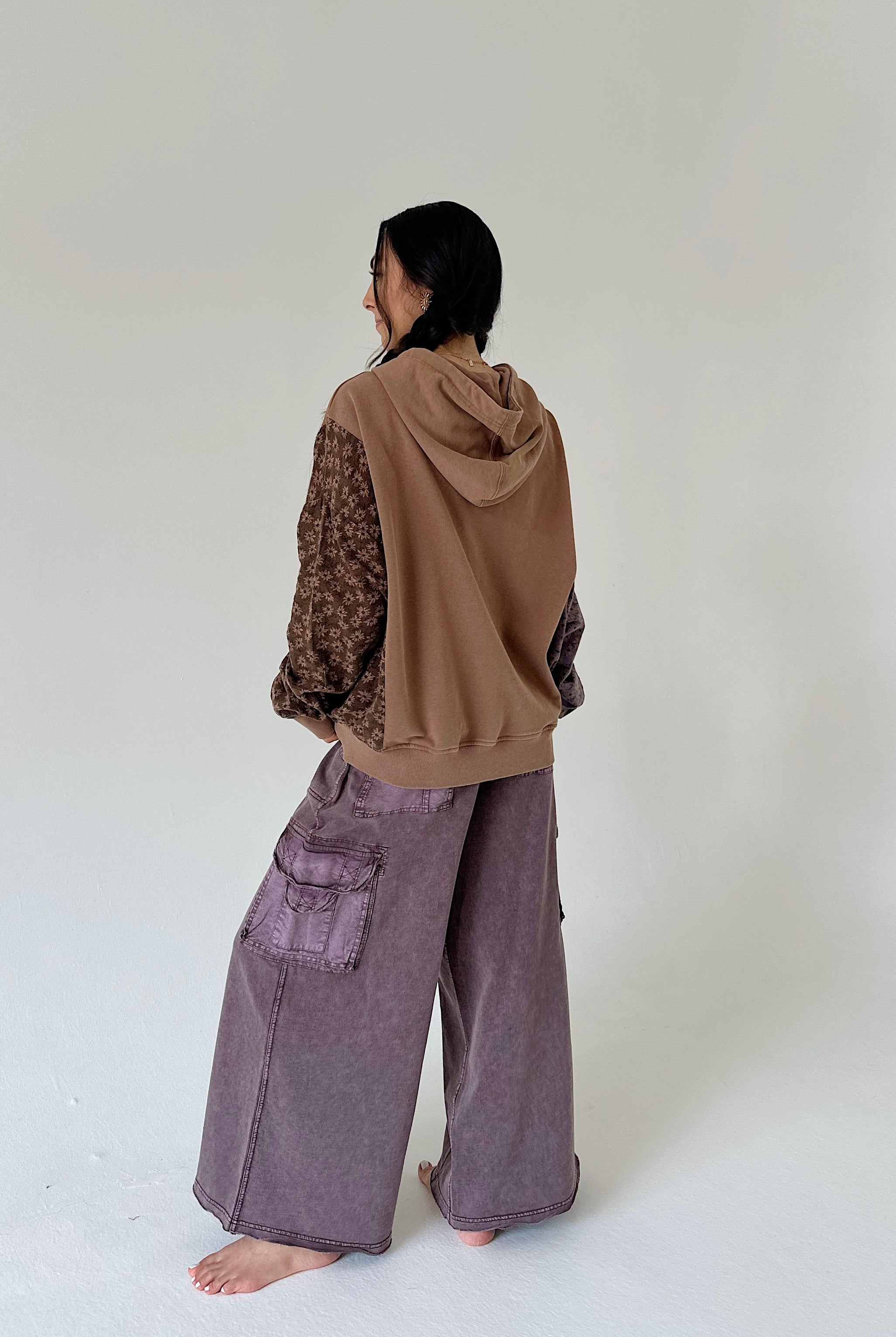 A person standing barefoot in a studio, shown from the back, wearing a brown hooded sweatshirt with patterned sleeves and wide-leg mauve-purple cargo pants.