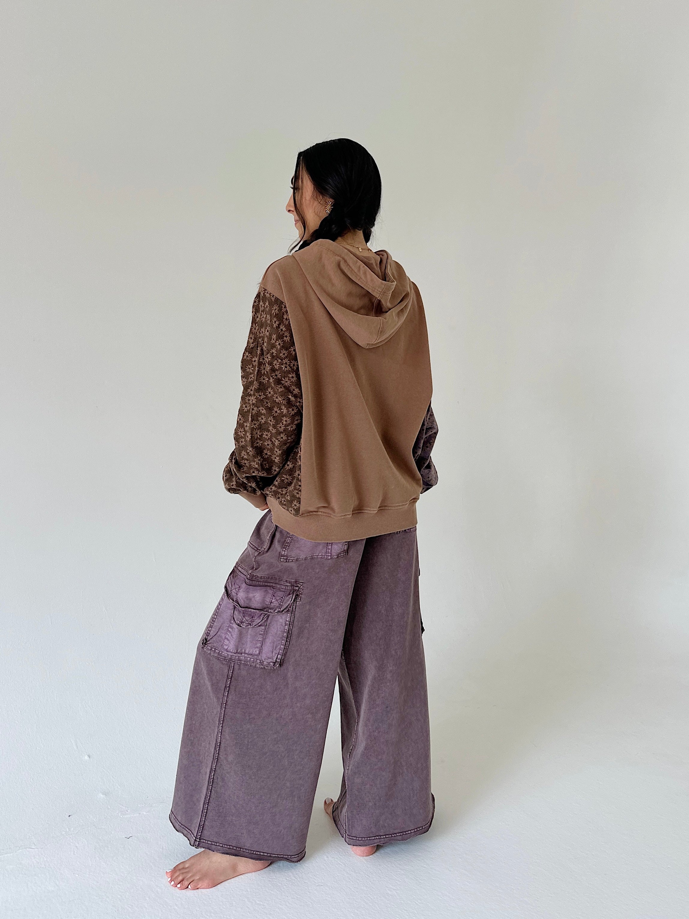 A person standing barefoot in a studio, shown from the back, wearing a brown hooded sweatshirt with patterned sleeves and wide-leg mauve-purple cargo pants.