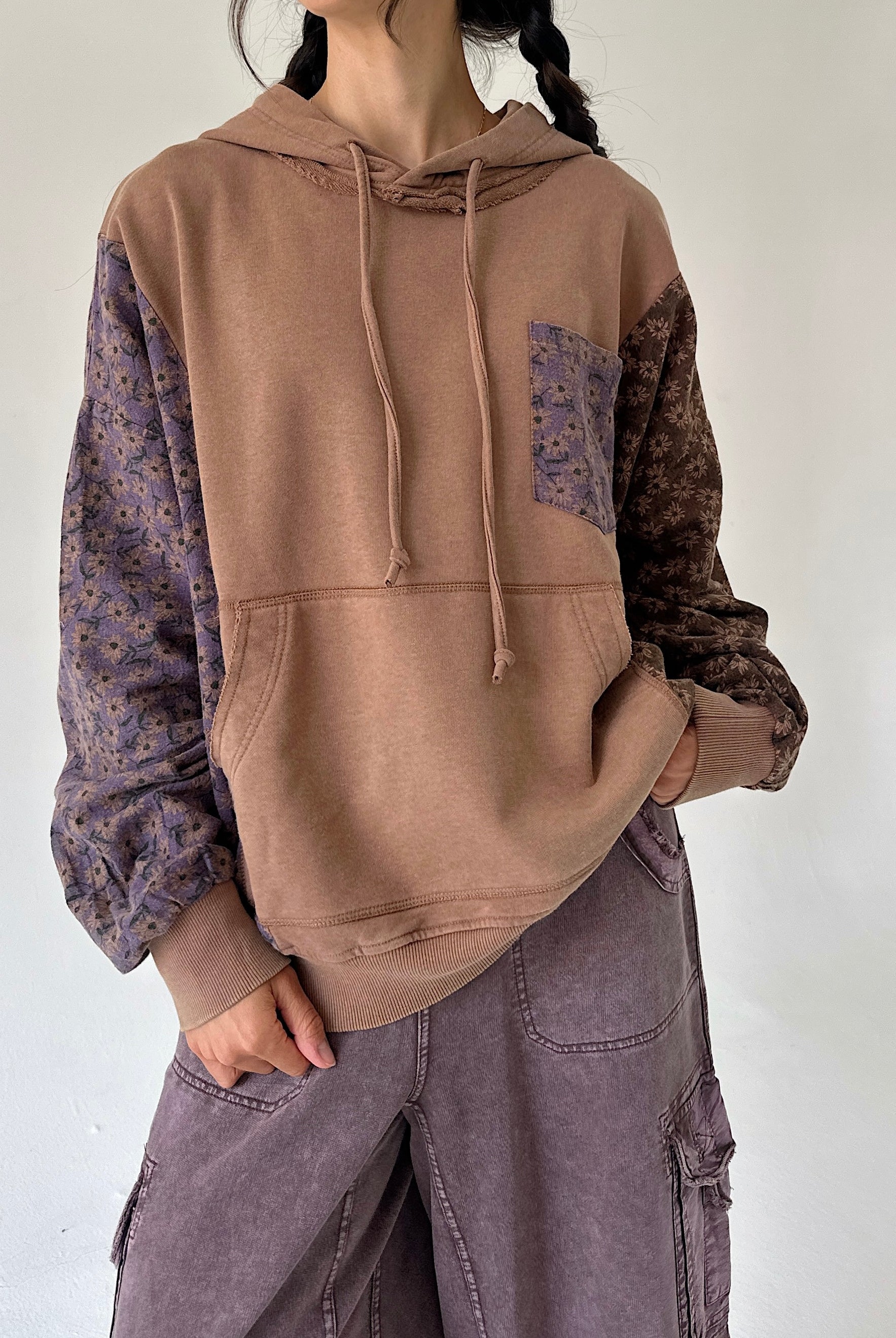 A person wearing a color-block hoodie with a solid tan front, purple floral patterned sleeves, and a dark brown floral chest pocket. The person is also wearing wide-leg mauve cargo pants against a plain off-white wall.