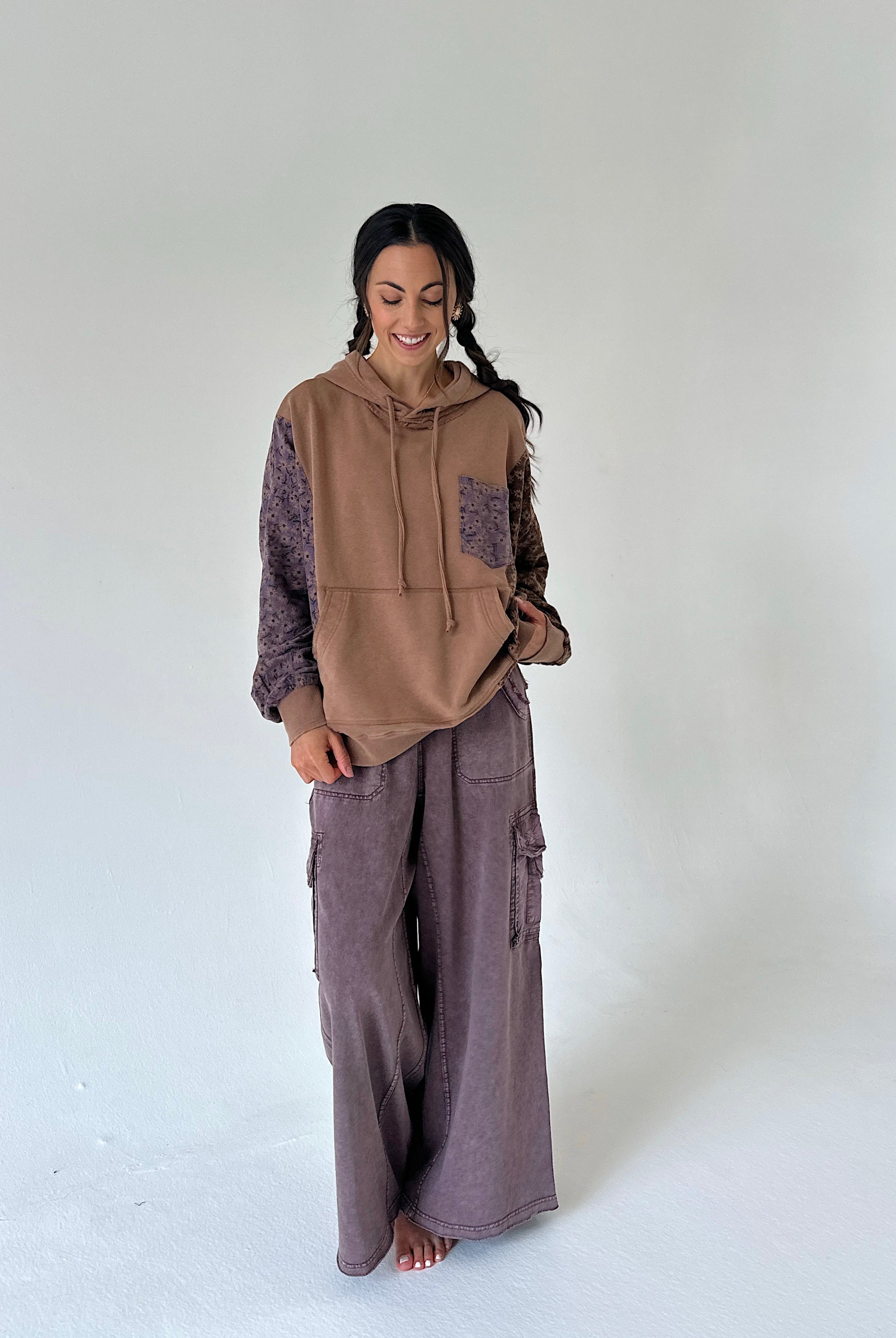 A woman stands barefoot against a plain light gray studio background, wearing a brown and mauve-purple patchwork hoodie with matching wide-leg cargo pants.