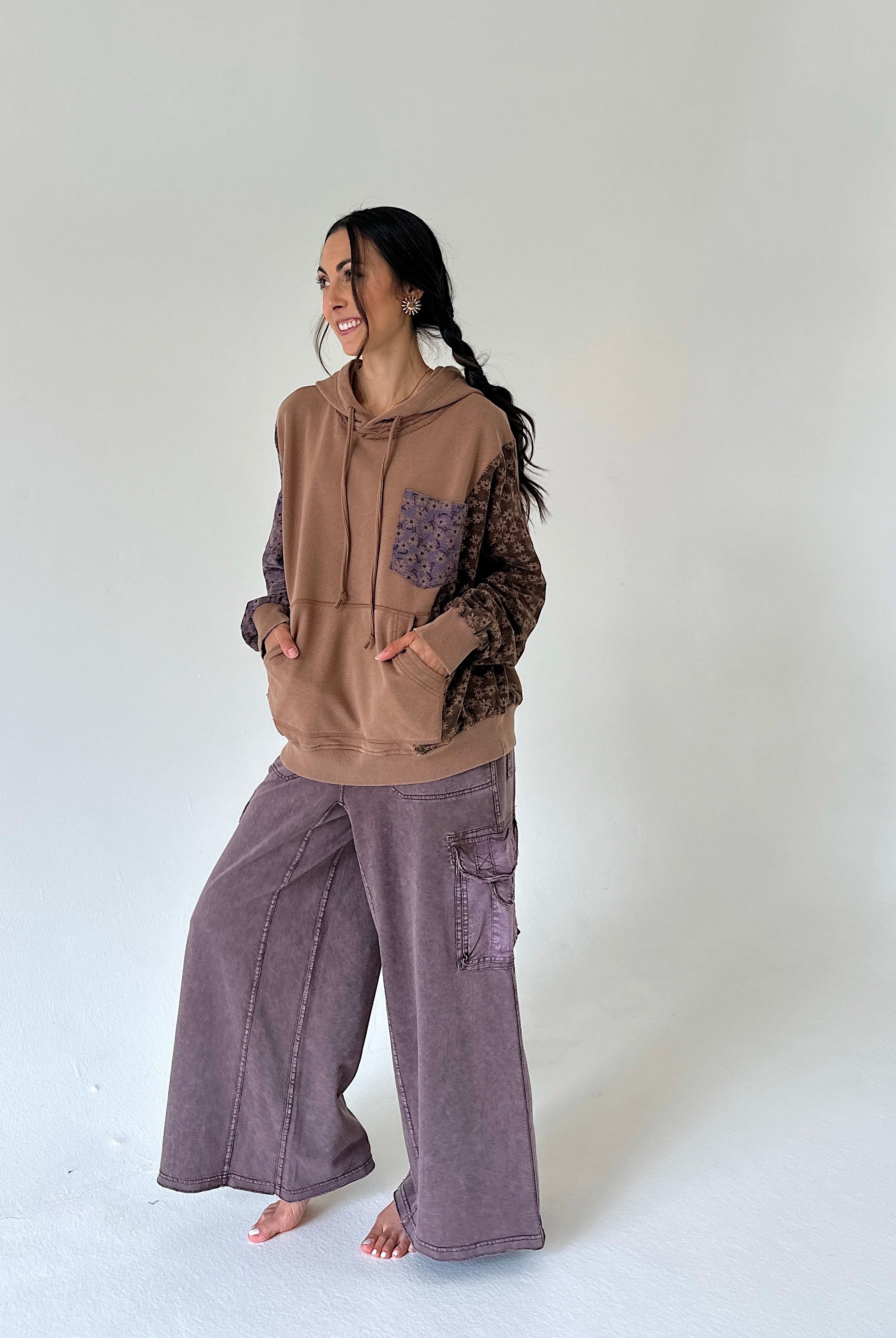 A woman modeling a brown and purple patchwork hoodie with patterned sleeves and wide-leg mauve-purple cargo pants, standing barefoot against a plain white studio background.