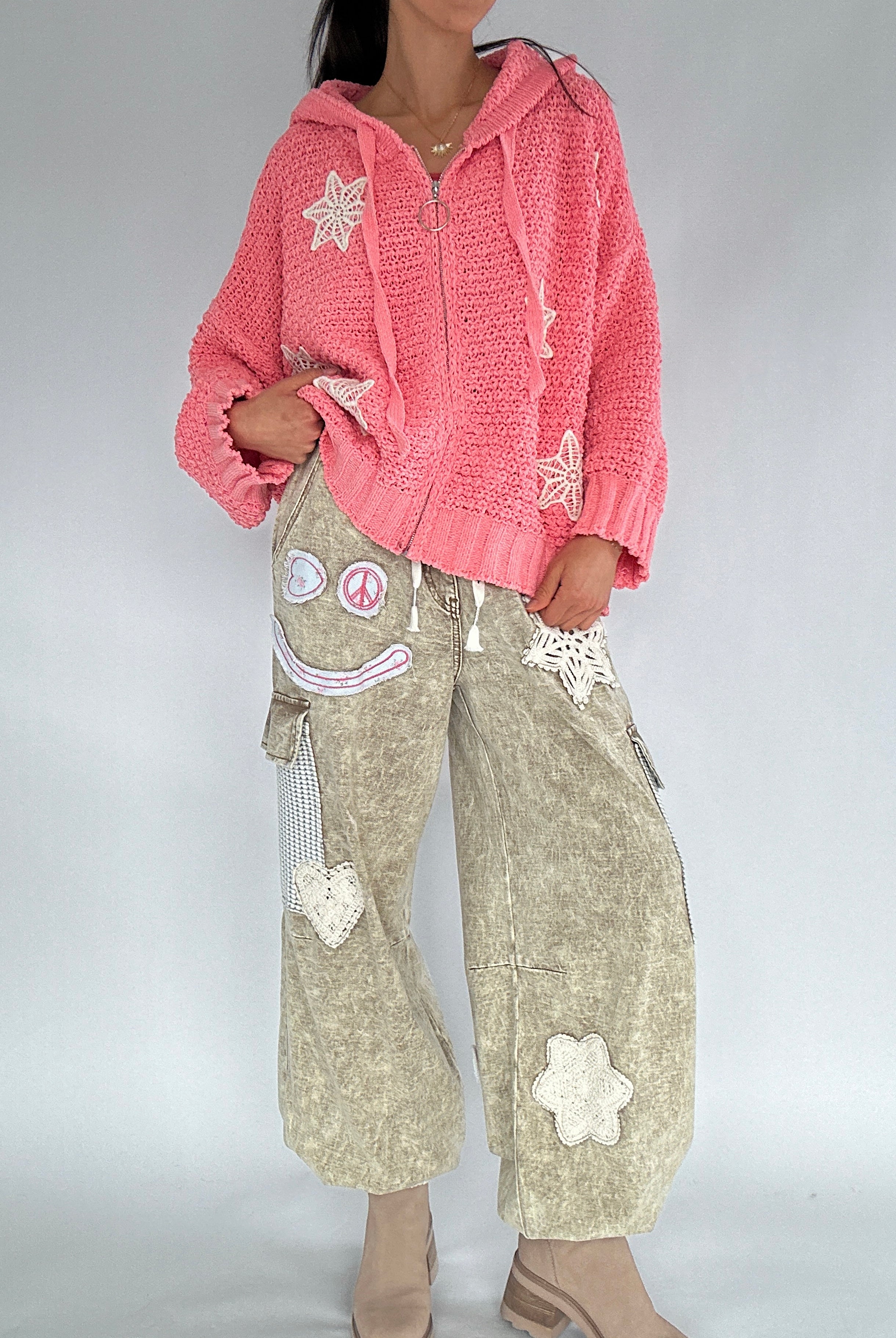 Person wearing a pink knitted hoodie and beige pants with floral patches on a plain background