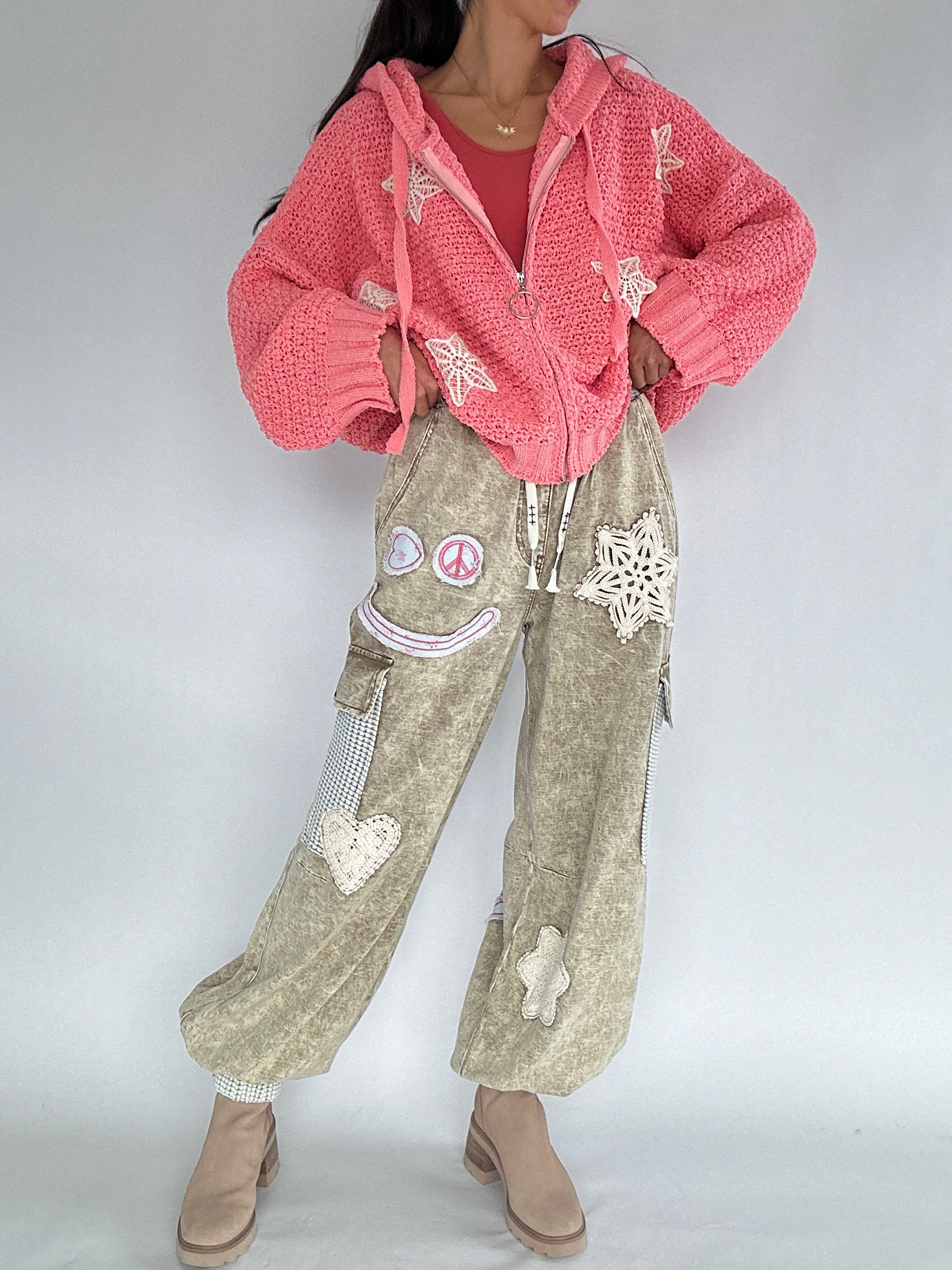 Person wearing a pink cardigan and beige cargo pants with decorative patches on a plain background
