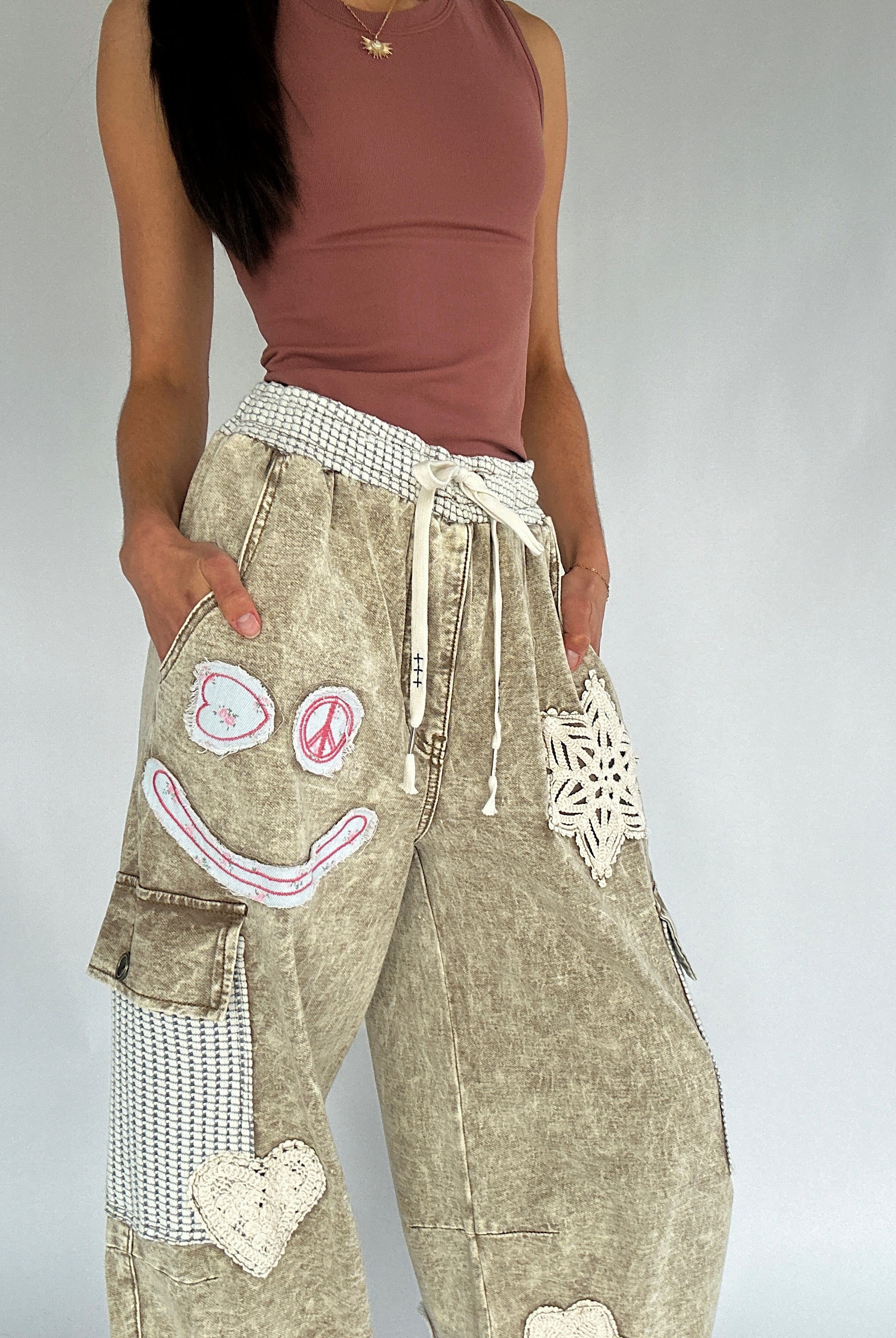 Person wearing a brown tank top and beige pants with decorative patches on a plain background