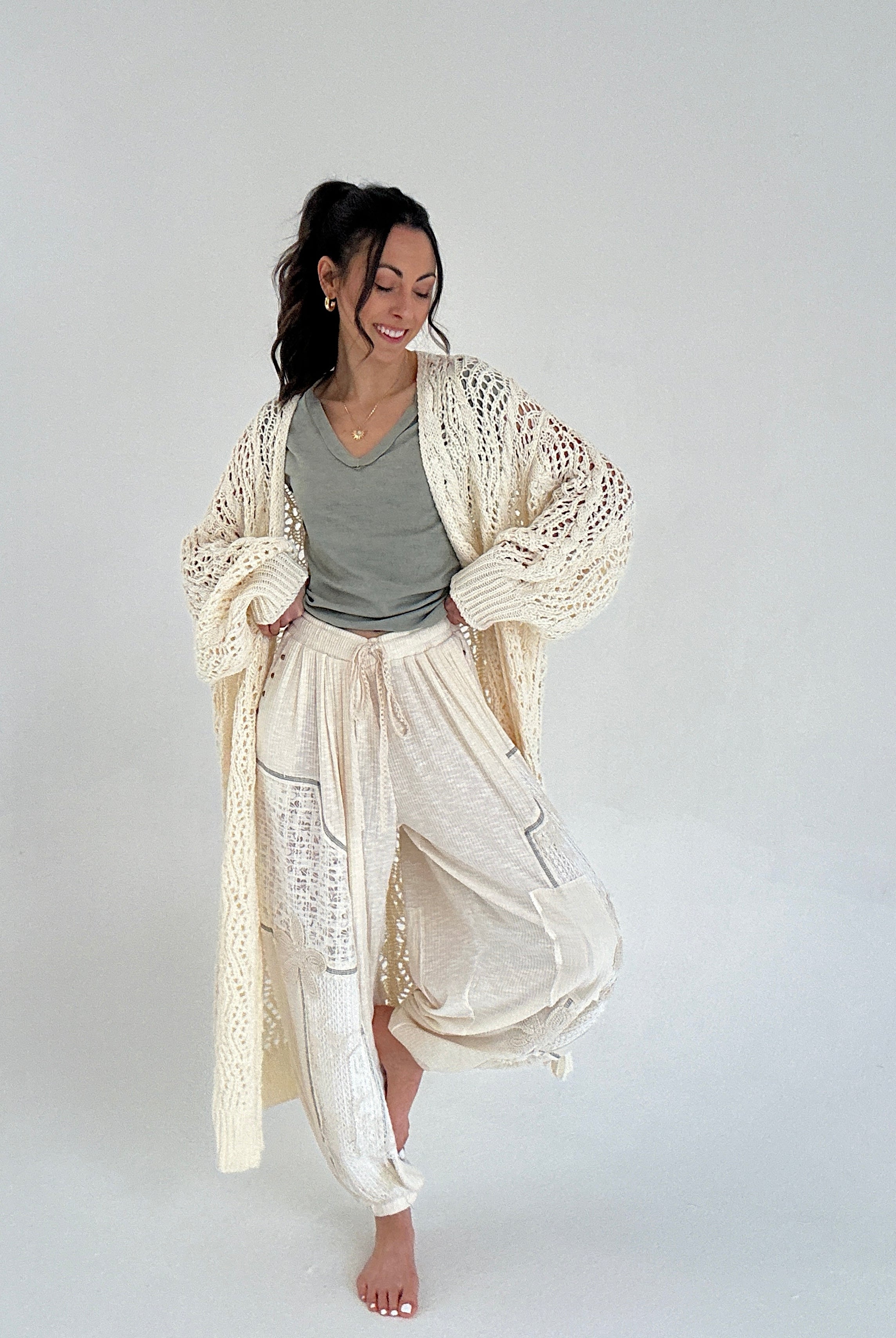 Woman wearing a long, light-colored cardigan over a green tee and cream jogger pants against a plain background