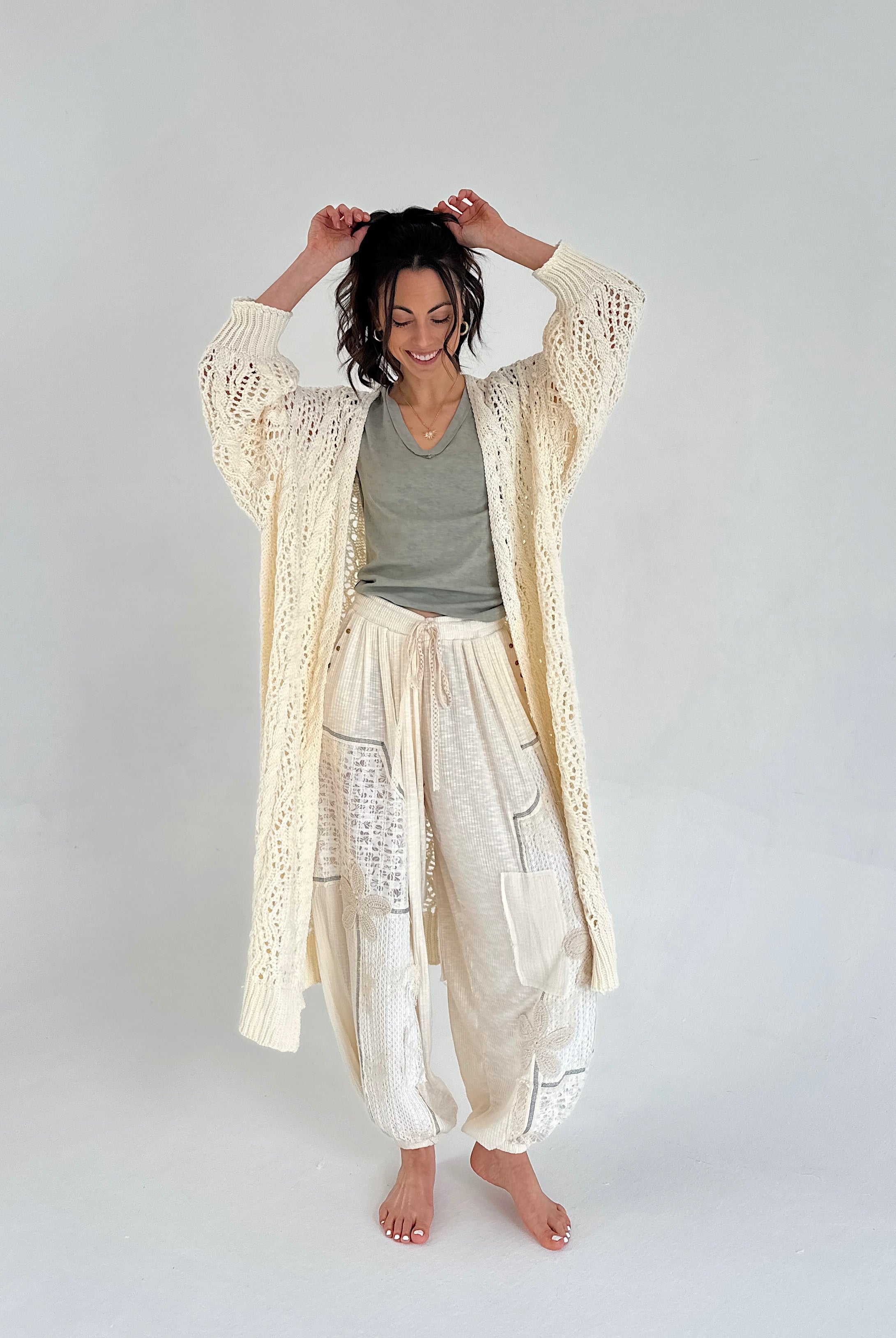 Woman wearing a long, light-colored cardigan over a gray top and white pants on a plain background