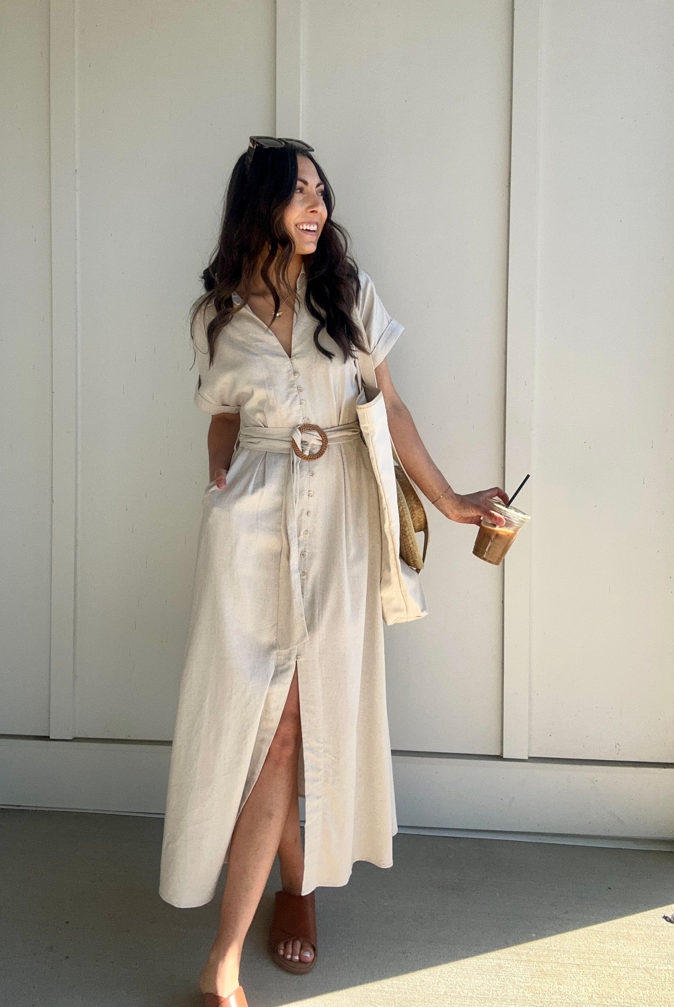 A woman wearing an oatmeal linen maxi dress with a waist belt, button front, lapel collar, front slit, and side pockets. She is holding a drink and wearing sandals.