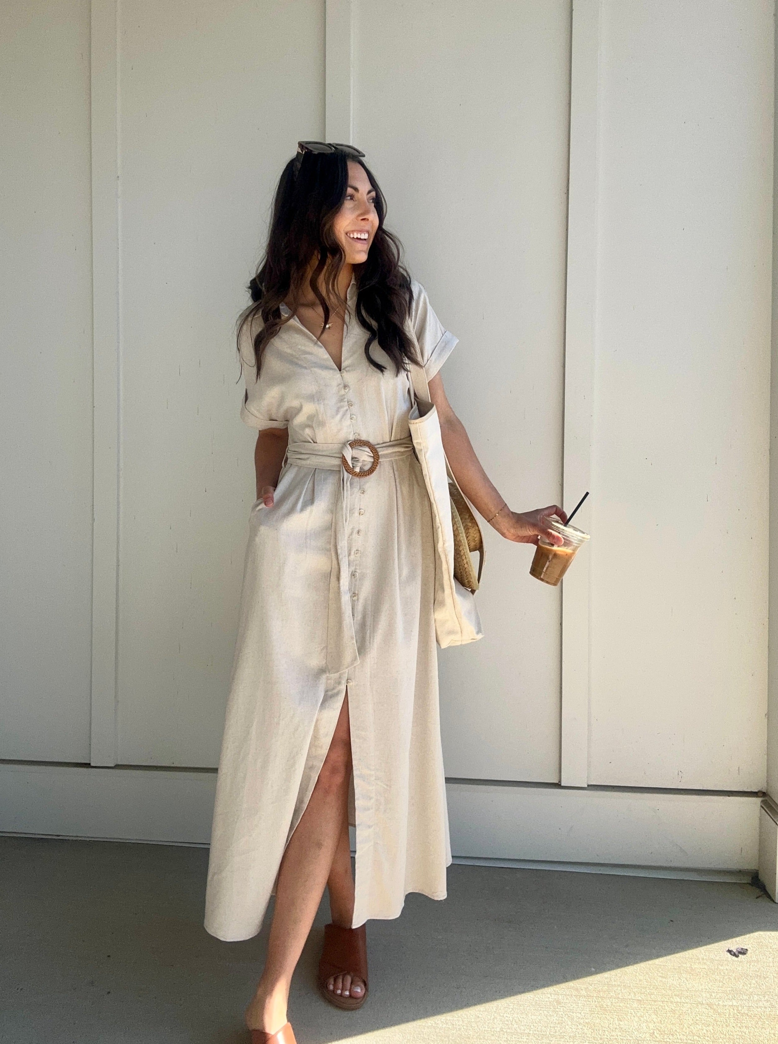 A woman wearing an oatmeal linen maxi dress with a waist belt, button front, lapel collar, front slit, and side pockets. She is holding a drink and wearing sandals.