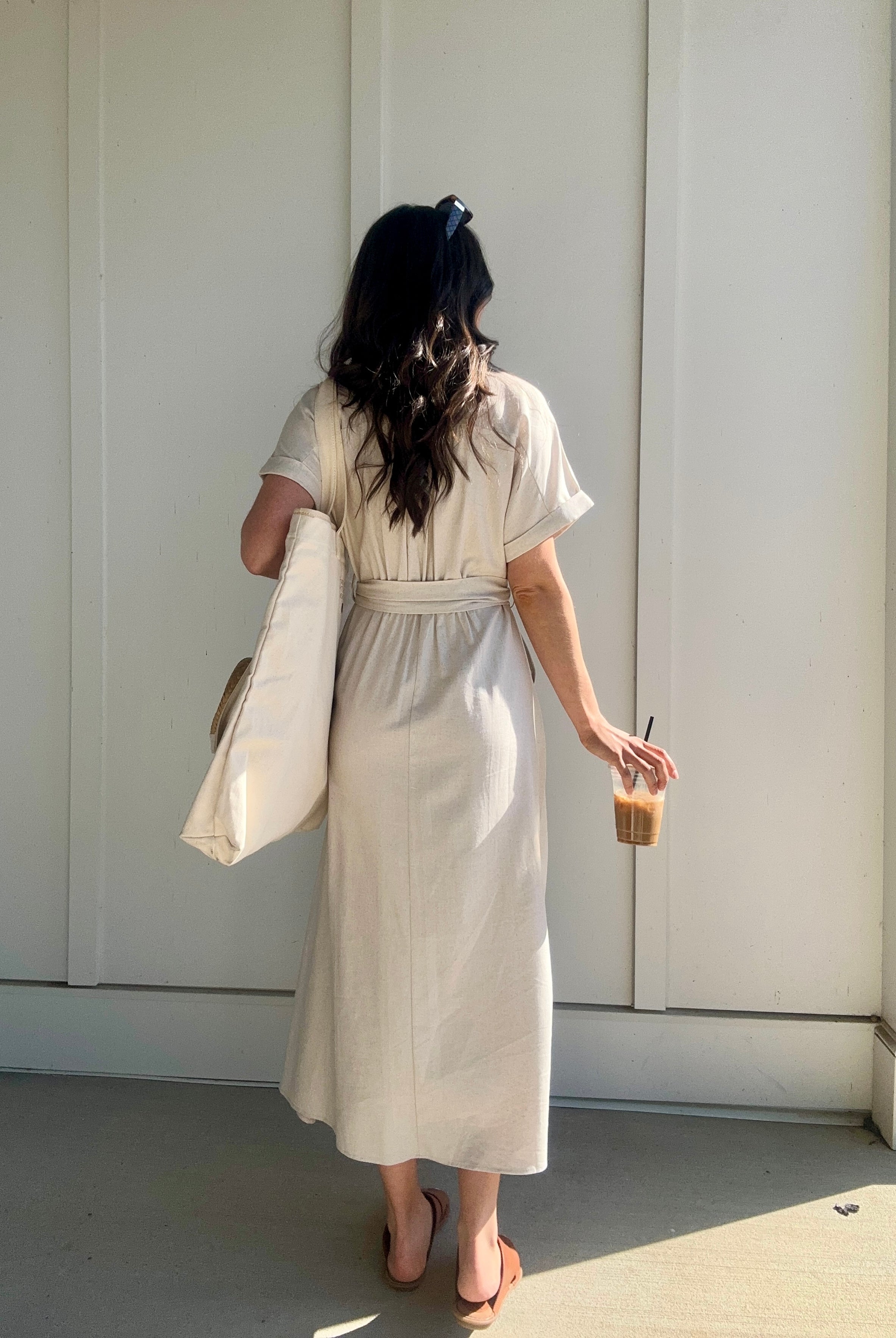 woman wearing oatmeal linen maxi dress from the back. she is holding a coffee and carrying a tote
