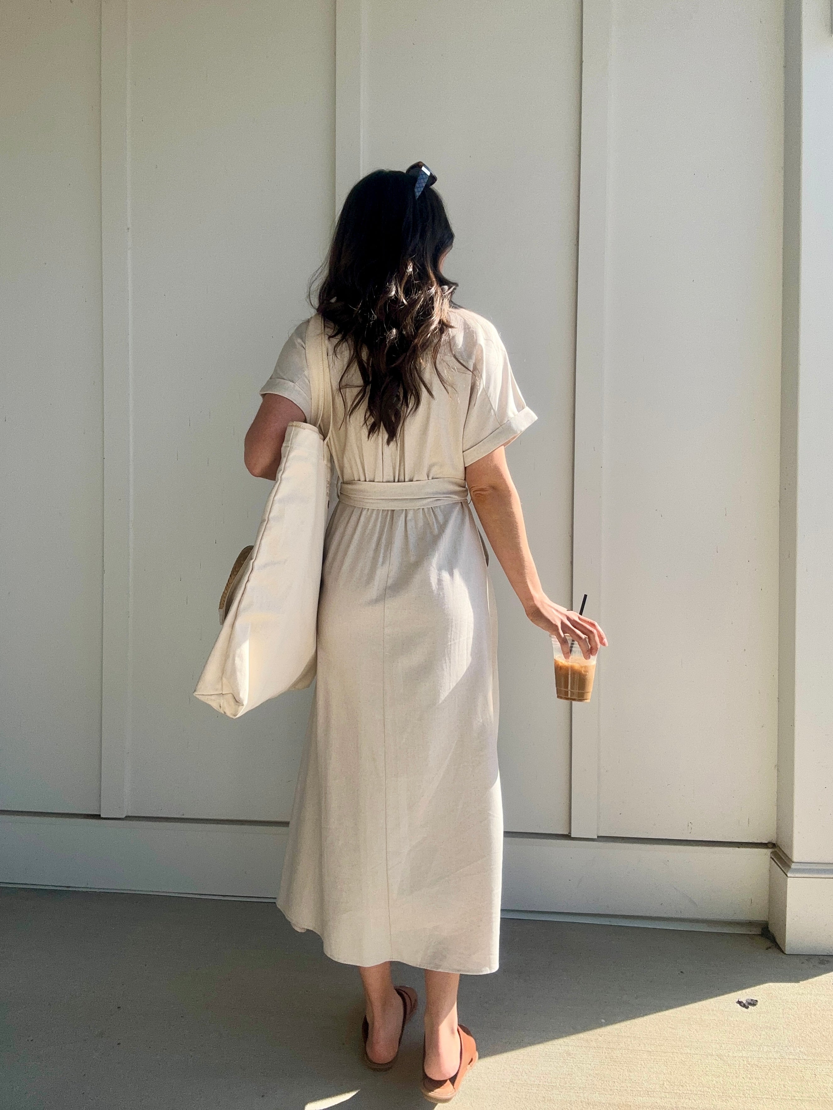 woman wearing oatmeal linen maxi dress from the back. she is holding a coffee and carrying a tote