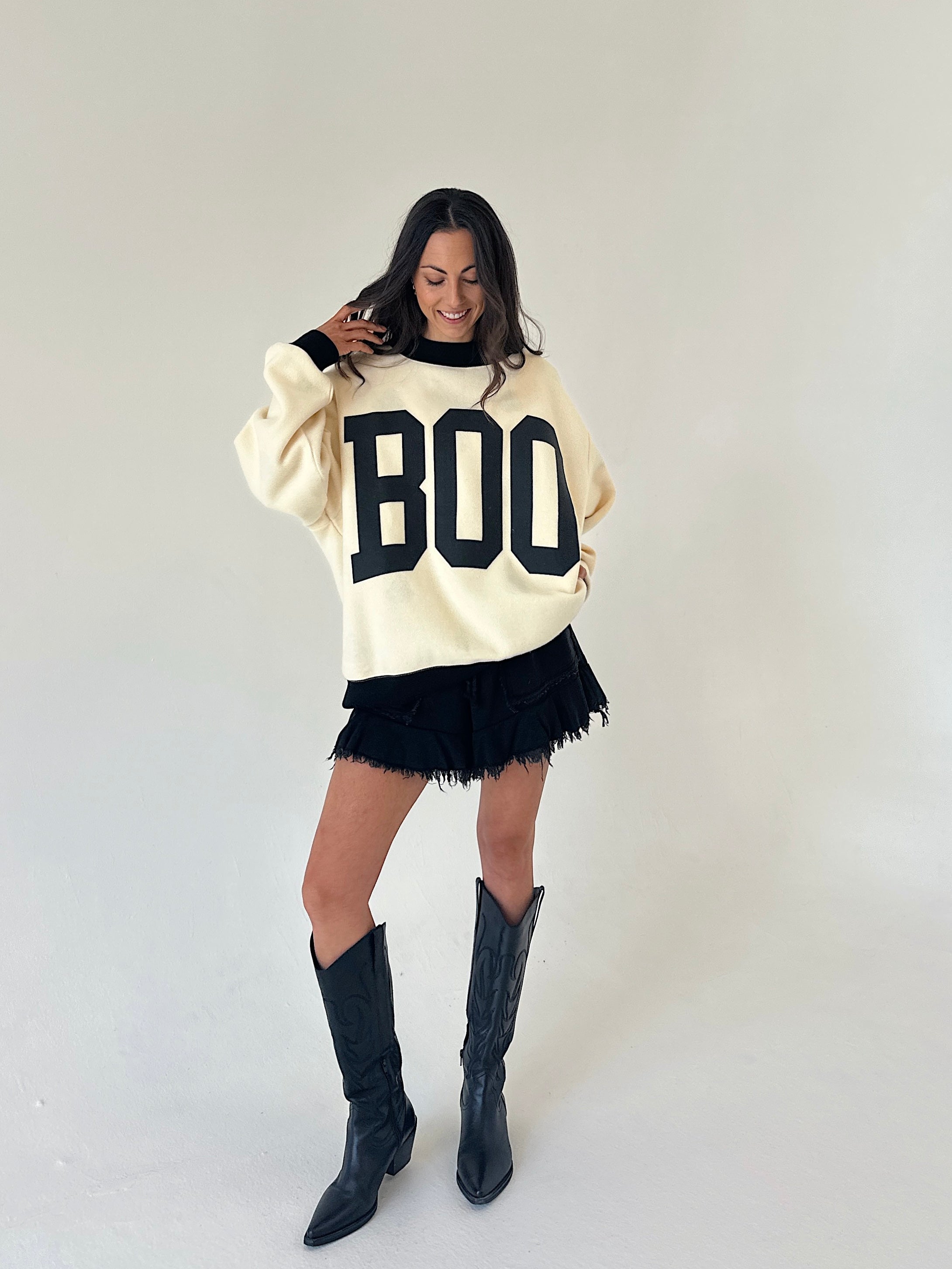 A woman modeling a cream-colored knit sweater with 'BOO' printed in black varsity letters, paired with a black fringed mini short and black knee-high boots, against a plain light gray studio background.