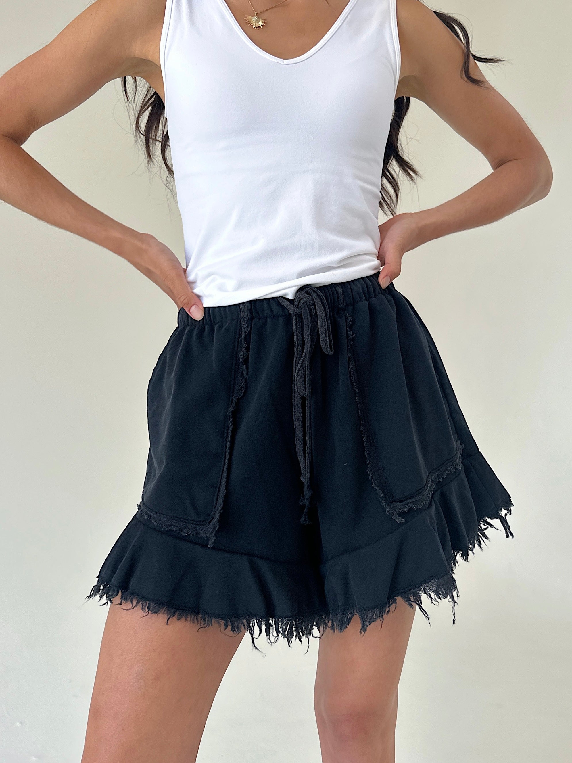 A model wearing black frayed terry shorts with a drawstring waist and a white tank top, standing against a plain off-white wall.