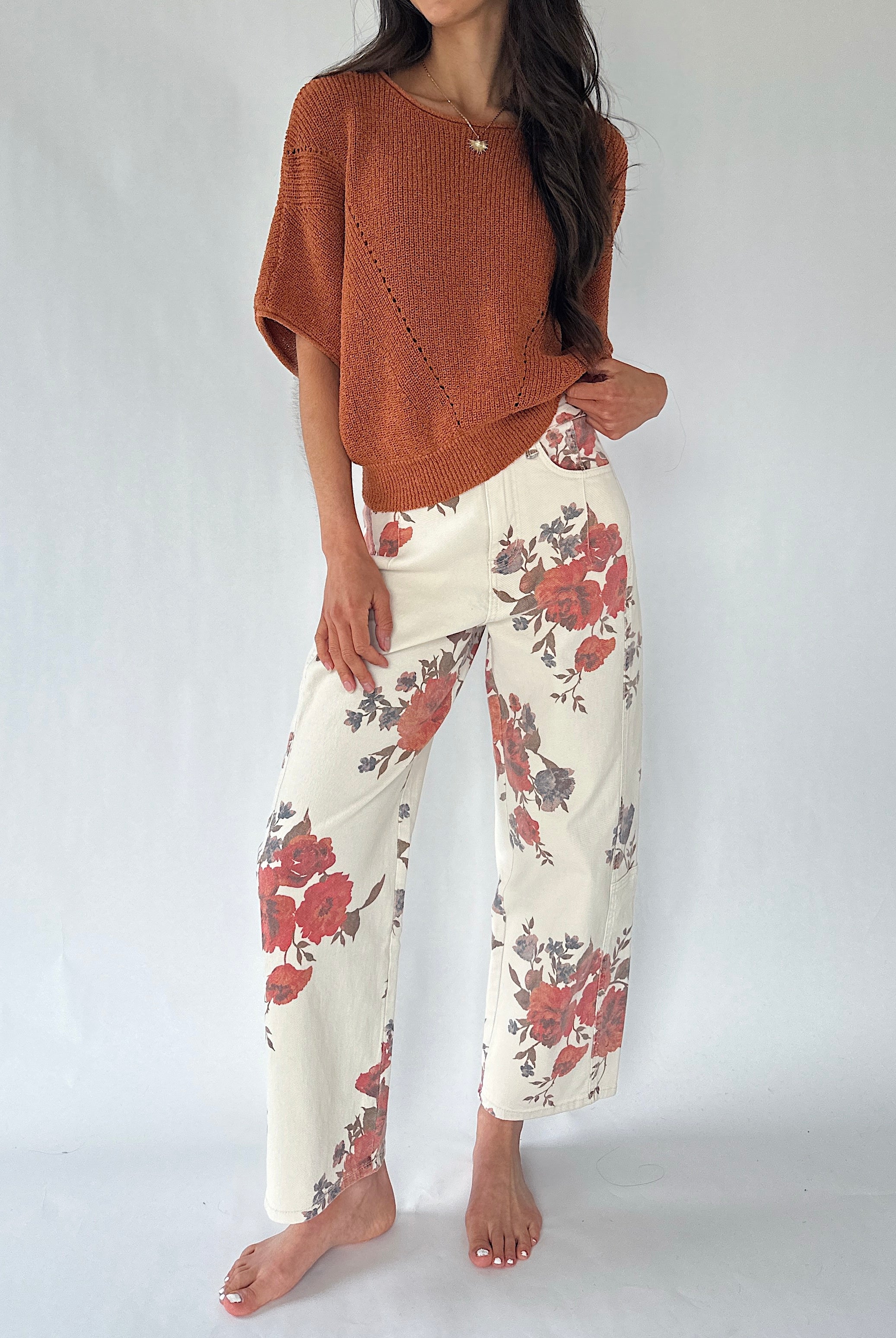 A woman modeling a rust-colored knit poncho sweater and cream wide-leg pants with a red and gray floral pattern, standing against a light gray studio background.