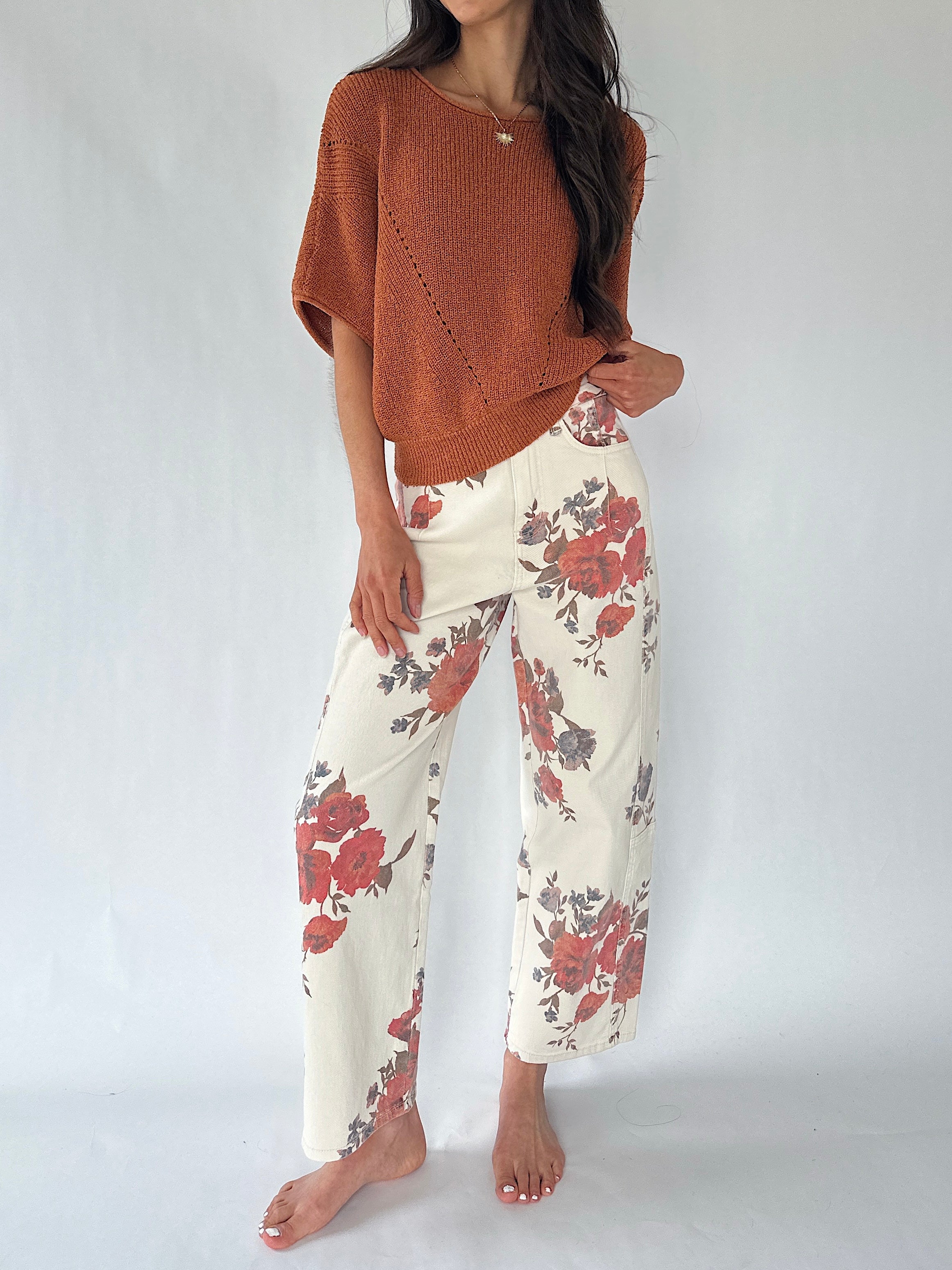 A woman modeling a rust-colored knit poncho sweater and cream wide-leg pants with a red and gray floral pattern, standing against a light gray studio background.