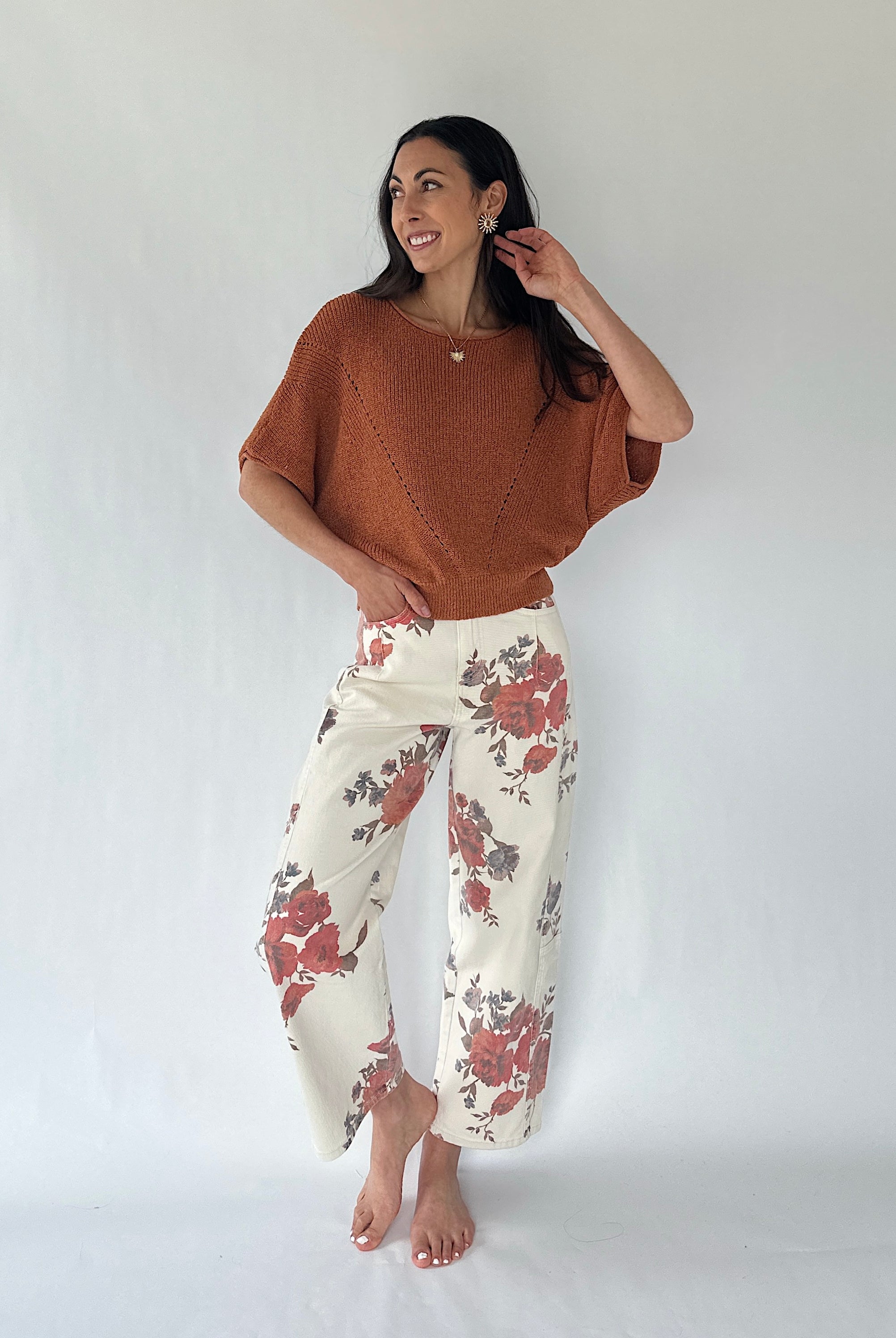 A woman modeling a rust-colored knit poncho top and cream wide-leg pants with a red and gray floral pattern, standing against a white studio backdrop.