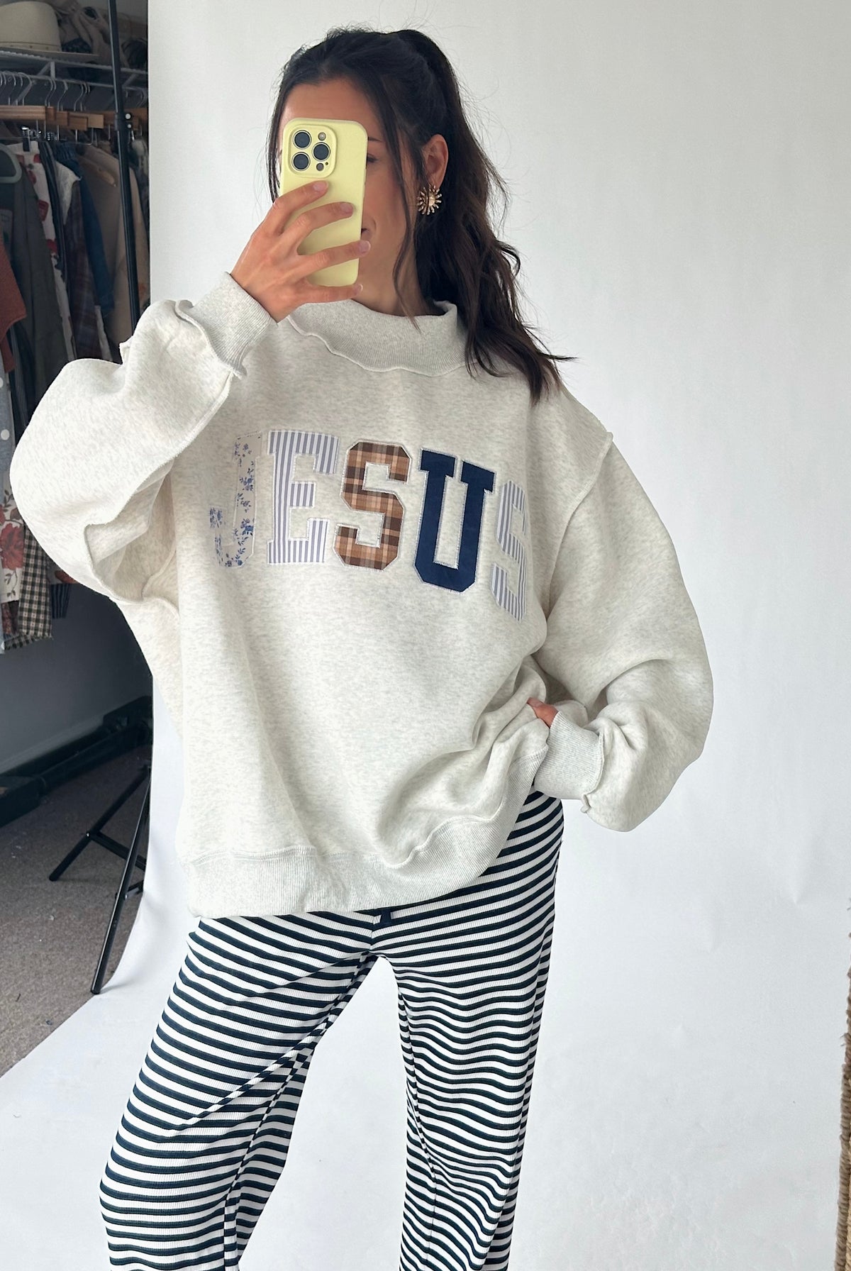 A woman takes a mirror selfie wearing a cream-colored sweatshirt with 'JESUS' printed across the chest in blue, brown, and white, paired with black and white striped pants.