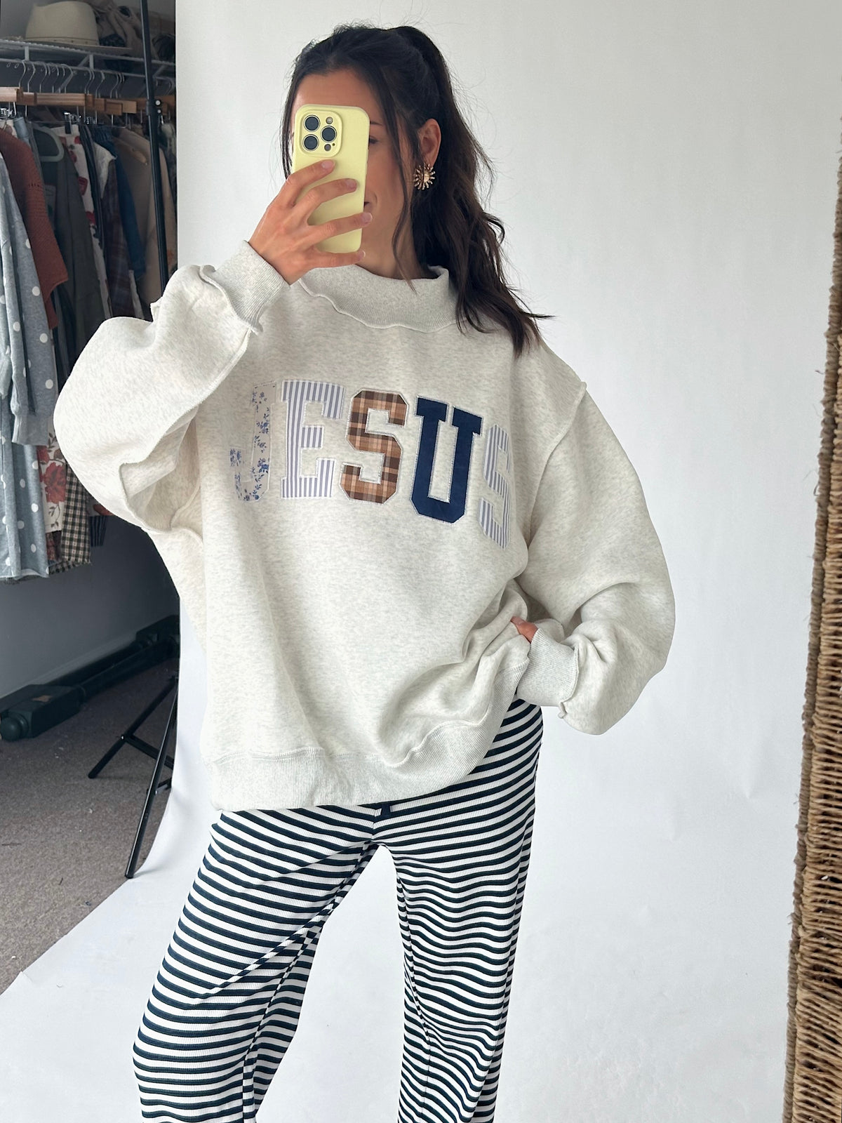 A woman takes a mirror selfie wearing a cream-colored sweatshirt with 'JESUS' printed across the chest in blue, brown, and white, paired with black and white striped pants.