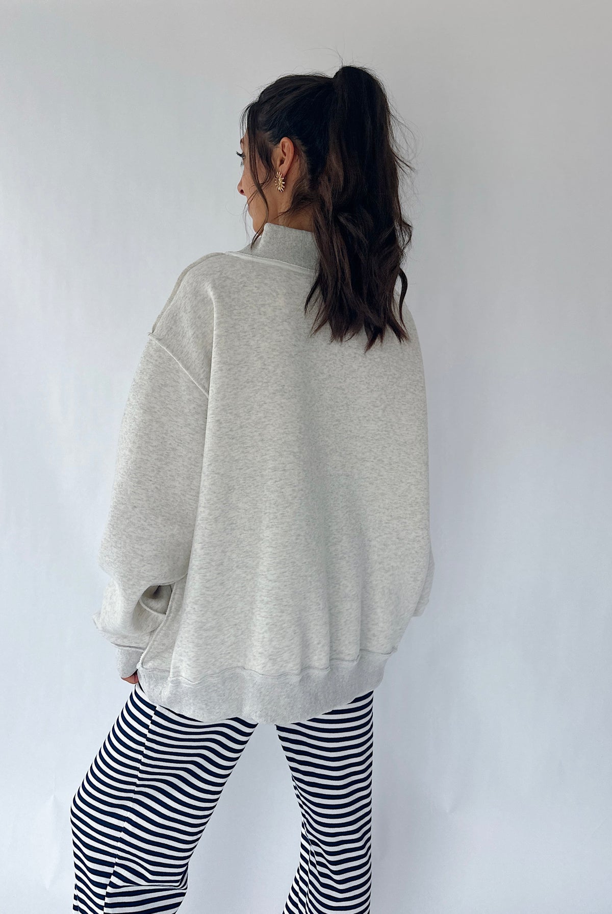 A woman shown from the back wearing an oversized light gray heather sweatshirt and black-and-white striped pants, standing against a plain light gray wall.